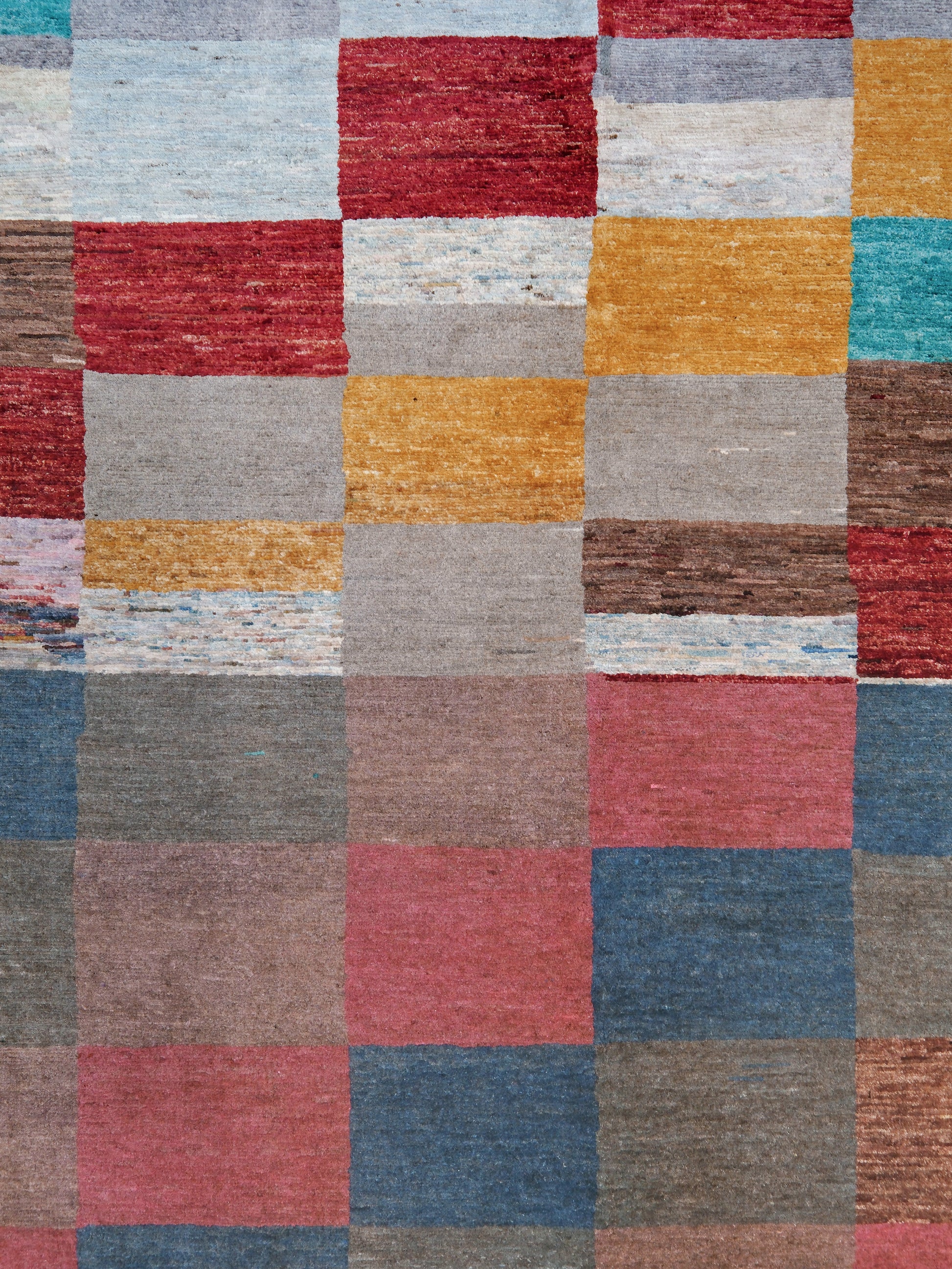 A close-up of a patchwork-style woven rug with a grid of rectangular blocks in muted red, mustard yellow, dusty pink, slate blue, taupe, brown, and off-white tones.