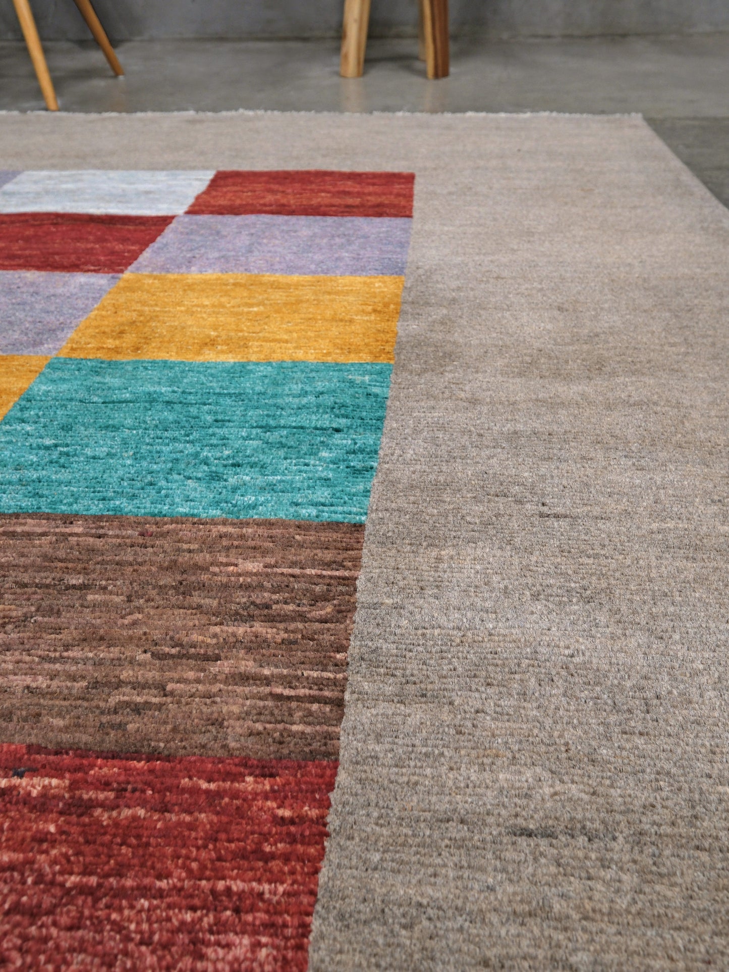 A close-up of a textured area rug with a large solid taupe-gray block on the right and a multicolor patchwork block on the left, featuring red, teal, yellow, lavender, and brown sections.