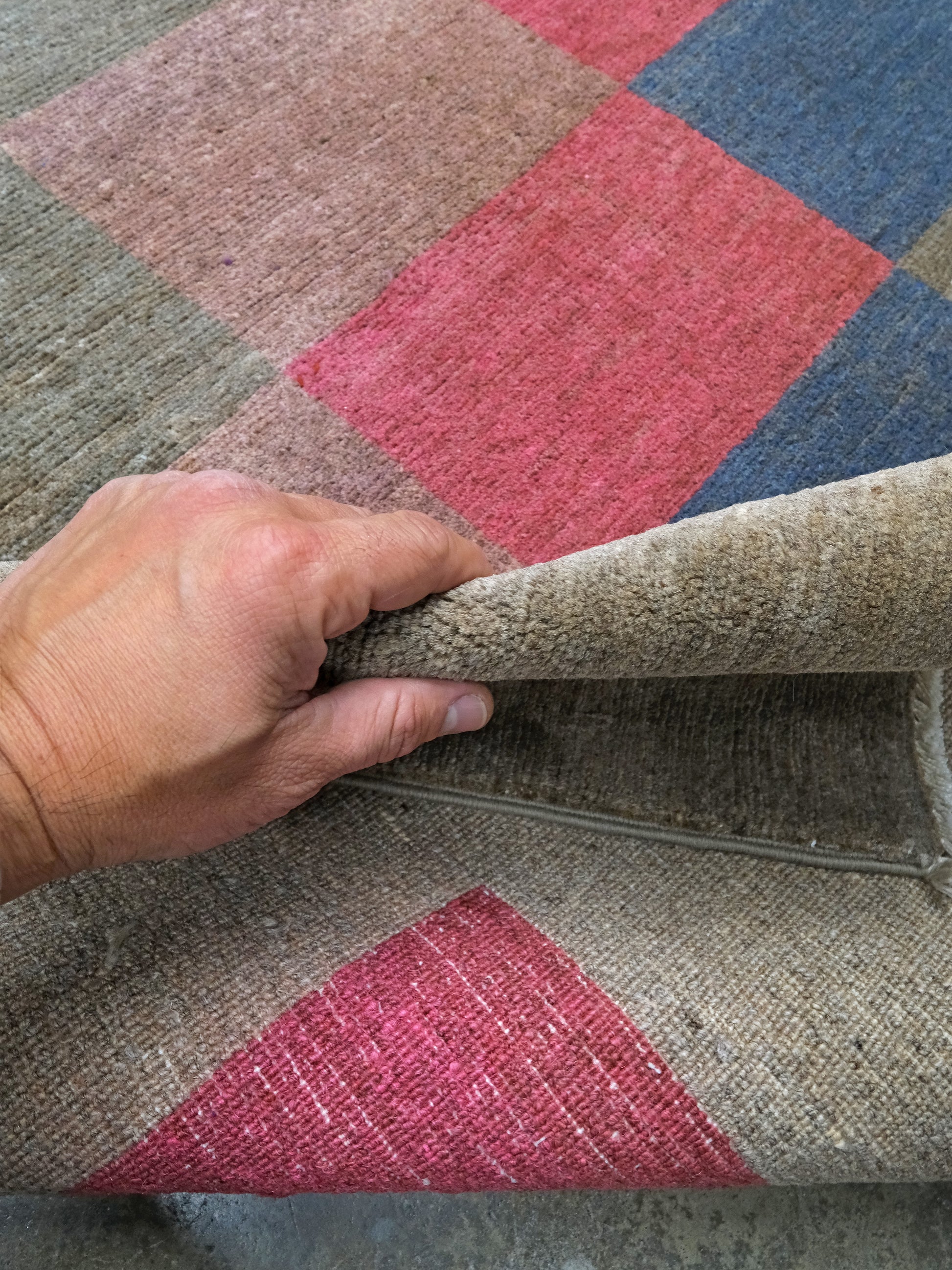 A close-up of a hand lifting the corner of a woven rug to show its texture and backing. The rug features a diamond pattern in muted beige, dusty pink, blue, and olive green.