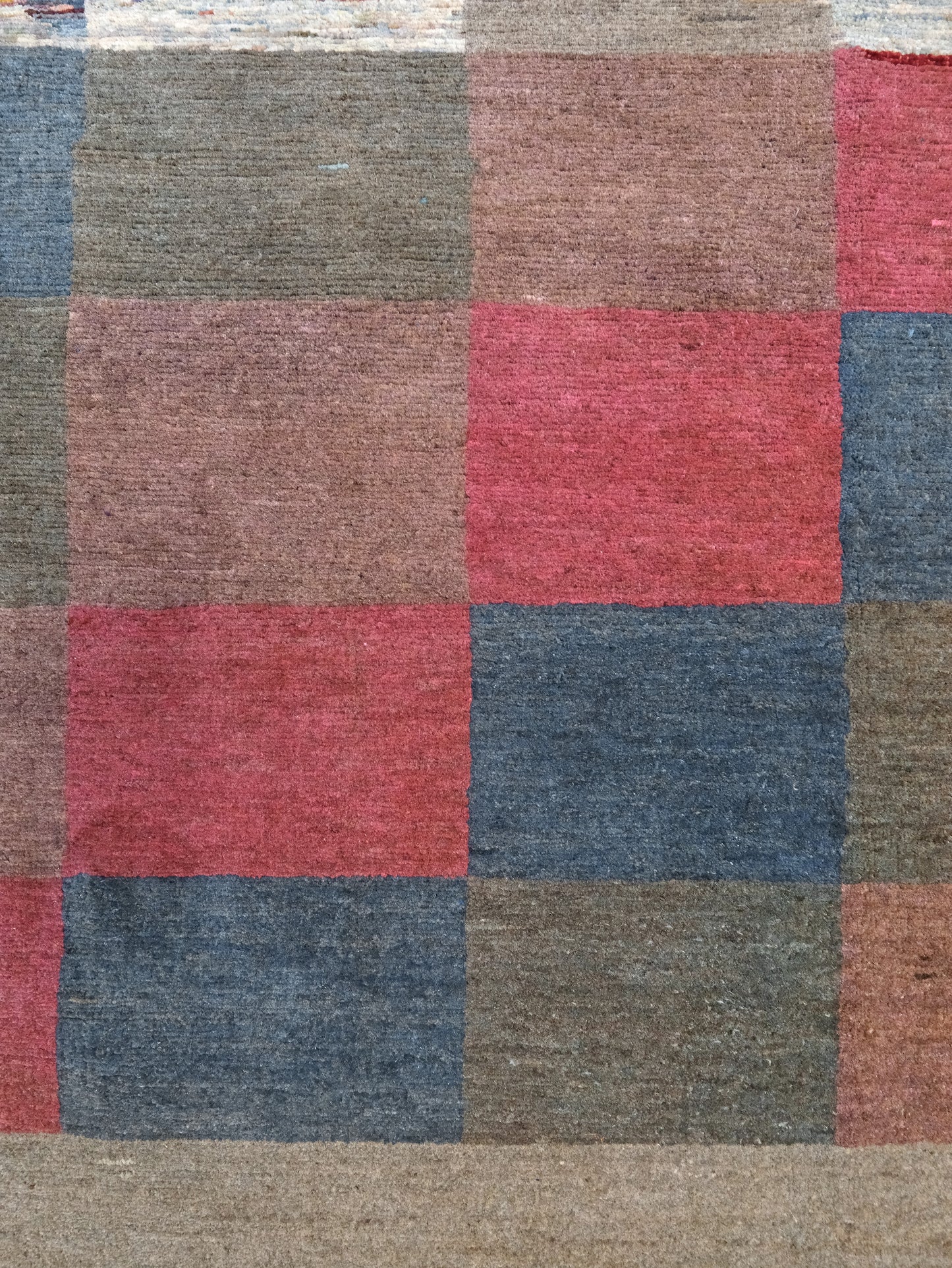 A close-up of a woven rug or textile with a patchwork grid pattern in muted tones of dusty red, slate blue, taupe, brown, and beige.