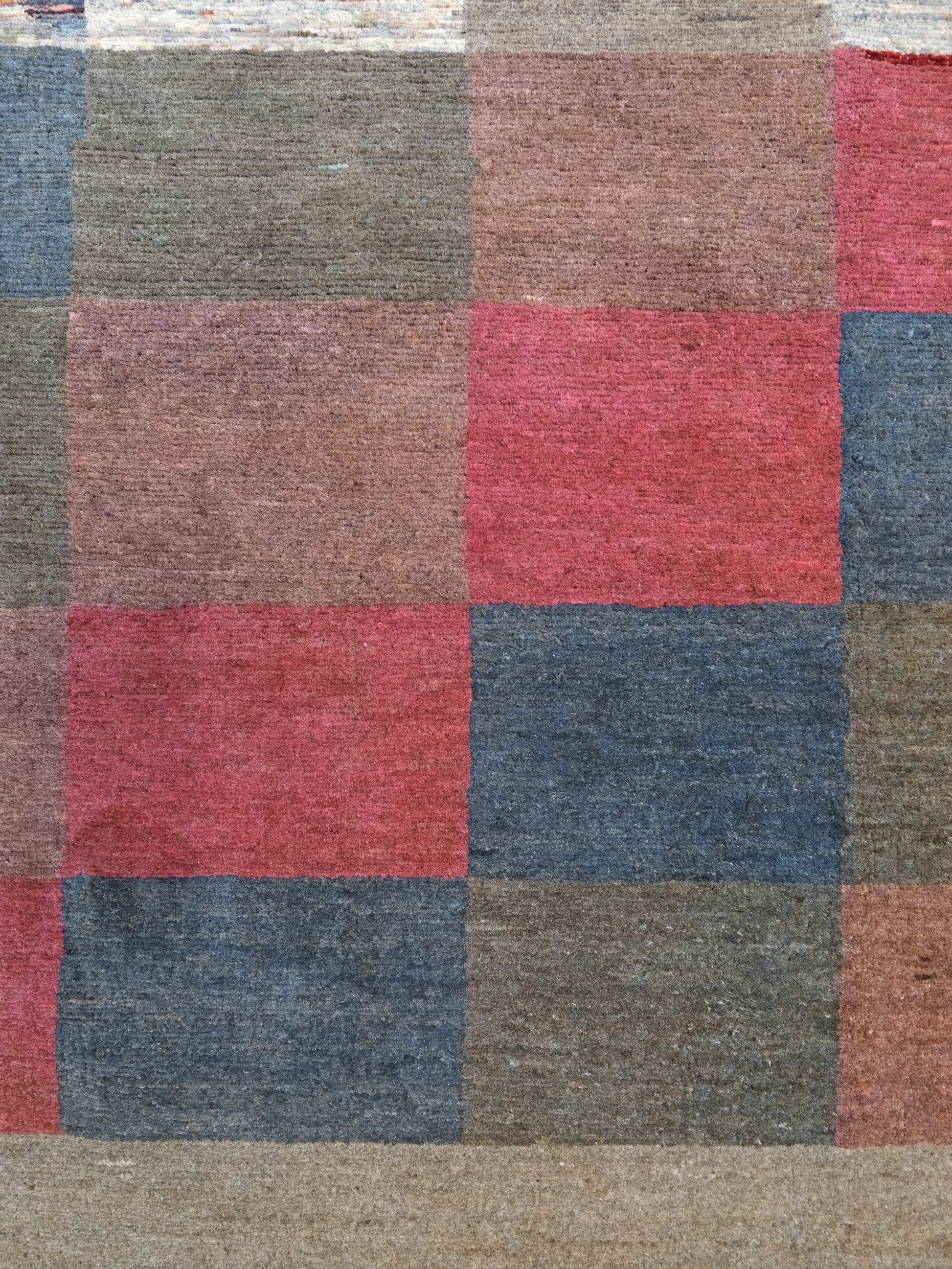 A close-up of a woven rug or textile with a patchwork grid pattern in muted tones of dusty red, slate blue, taupe, brown, and beige.