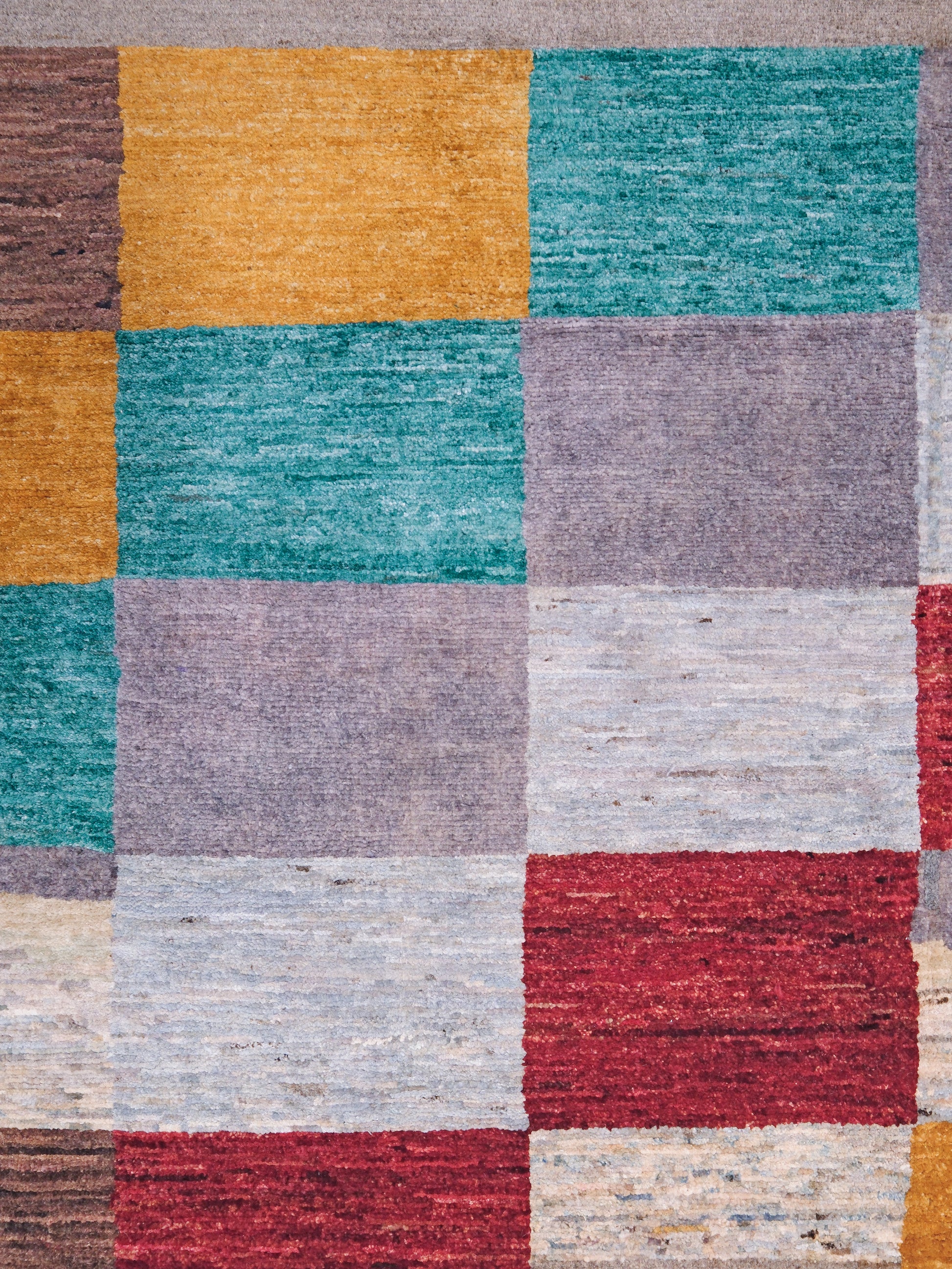 A close-up of a textured patchwork rug featuring a grid of square blocks in muted teal, mustard yellow, lavender, light gray, red, and cream.