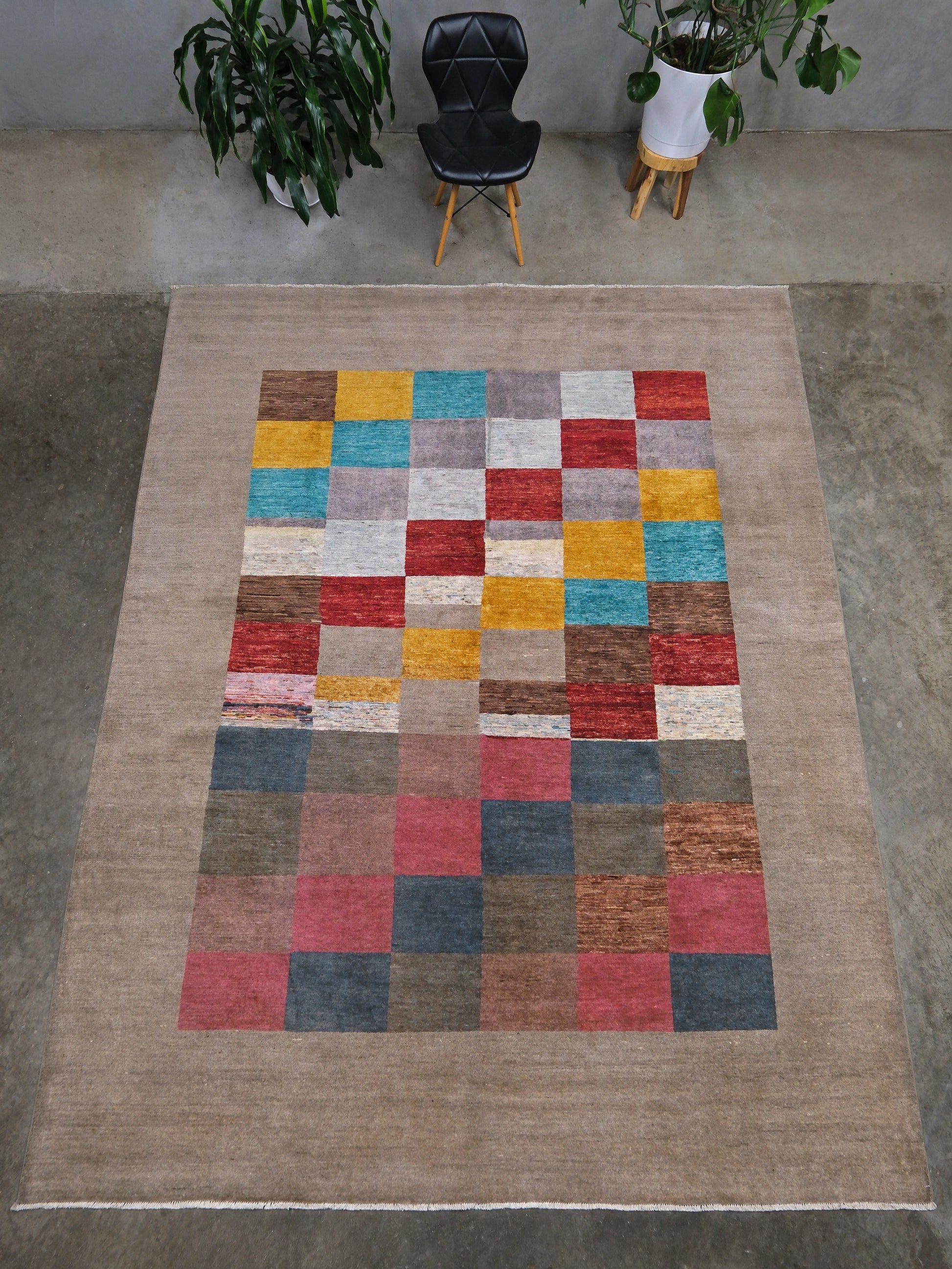 A handmade modern area rug laid on a concrete floor, featuring a large beige border and a central patchwork design made of multicolored squares in red, blue, yellow, gray, and brown. A black chair and potted plants are visible in the background.