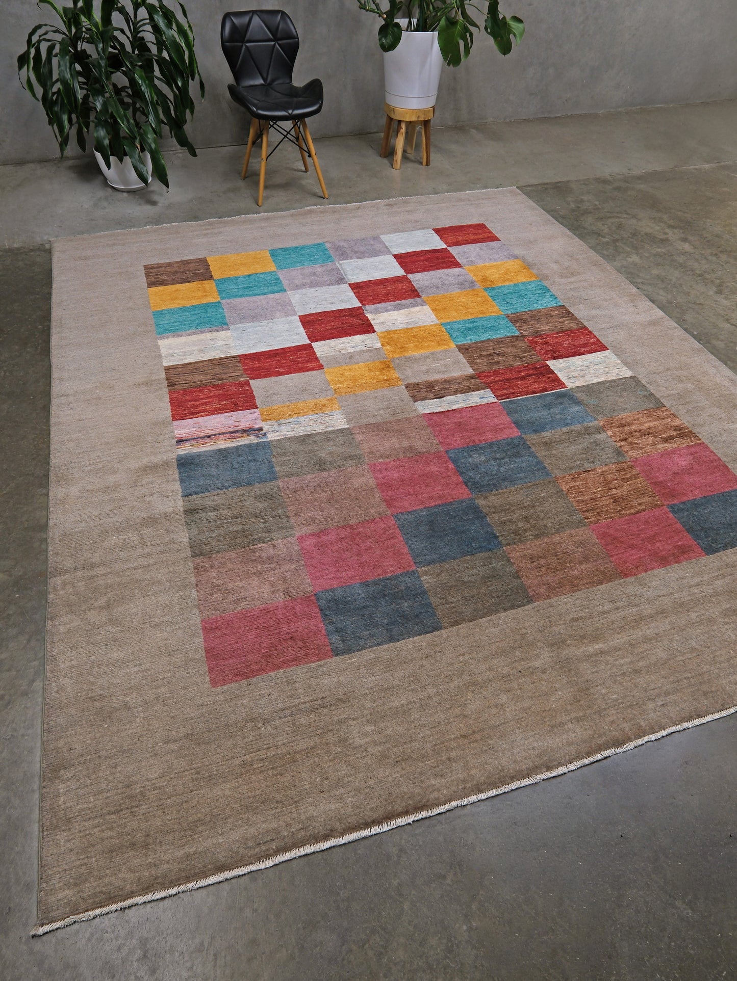 A large rectangular area rug with a beige base and a central patchwork-style grid of multicolored squares in red, blue, yellow, pink, and gray, shown on a concrete floor in a styled room with plants and a chair.