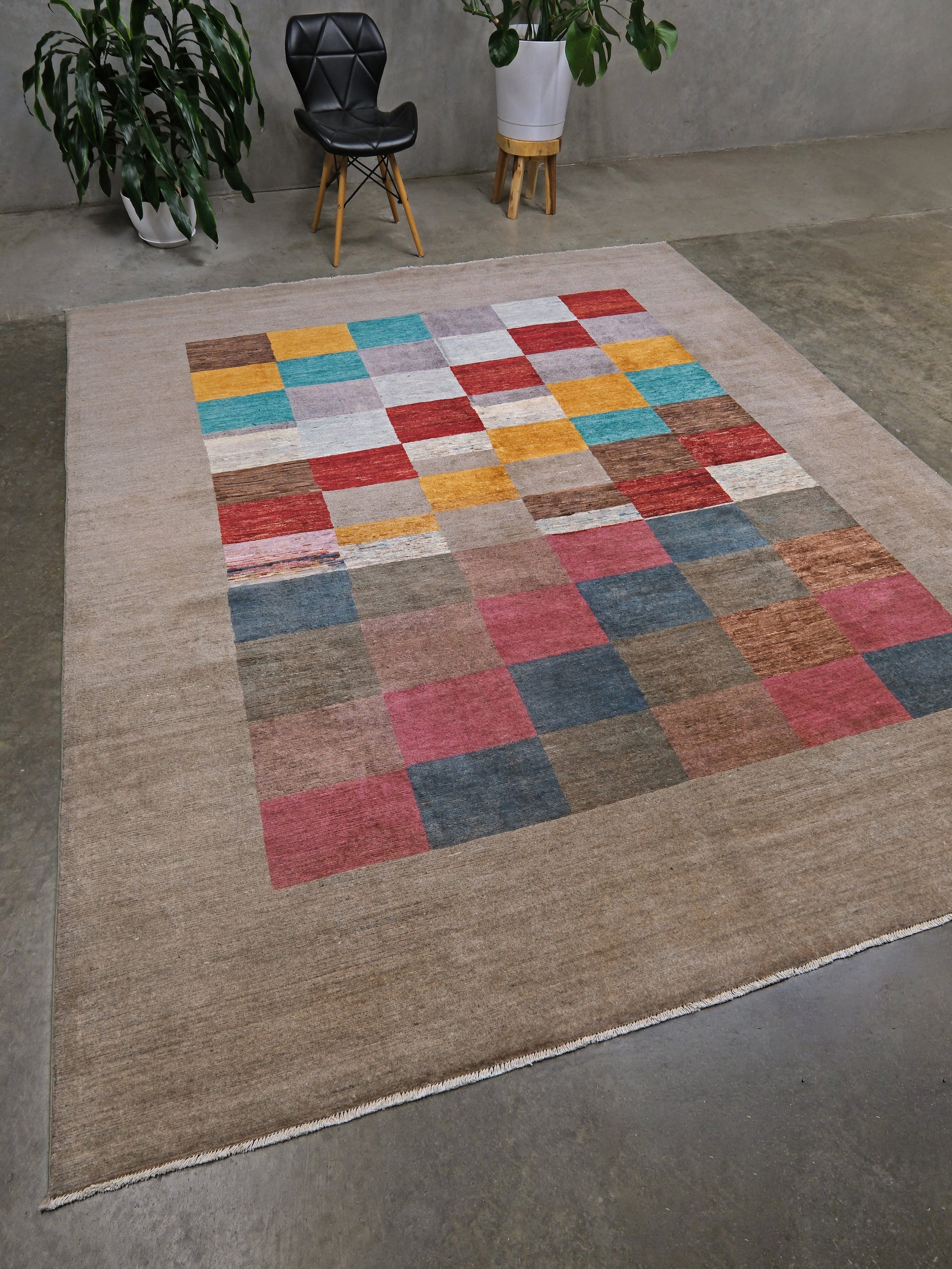 A large rectangular area rug with a beige base and a central patchwork-style grid of multicolored squares in red, blue, yellow, pink, and gray, shown on a concrete floor in a styled room with plants and a chair.