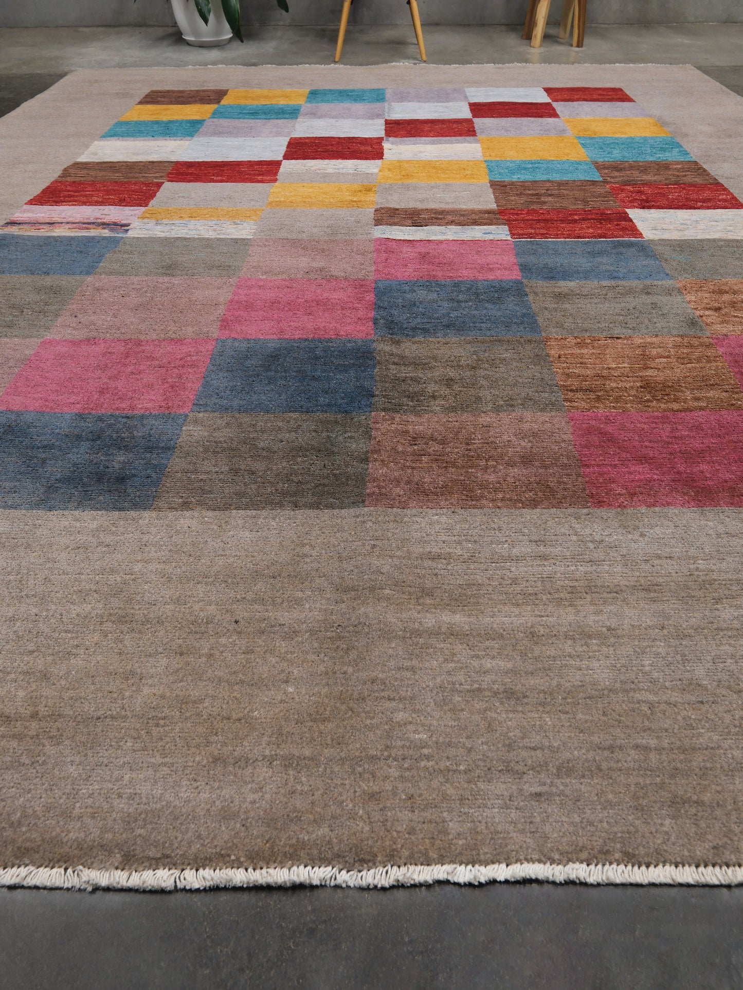 A large rectangular hanmade area rug with a patchwork design, featuring a wide taupe-brown field at the bottom that transitions into a grid of multicolored square blocks above. The rug is shown laid out on a dark gray concrete floor, with the legs of two chairs partially visible at the top edge.