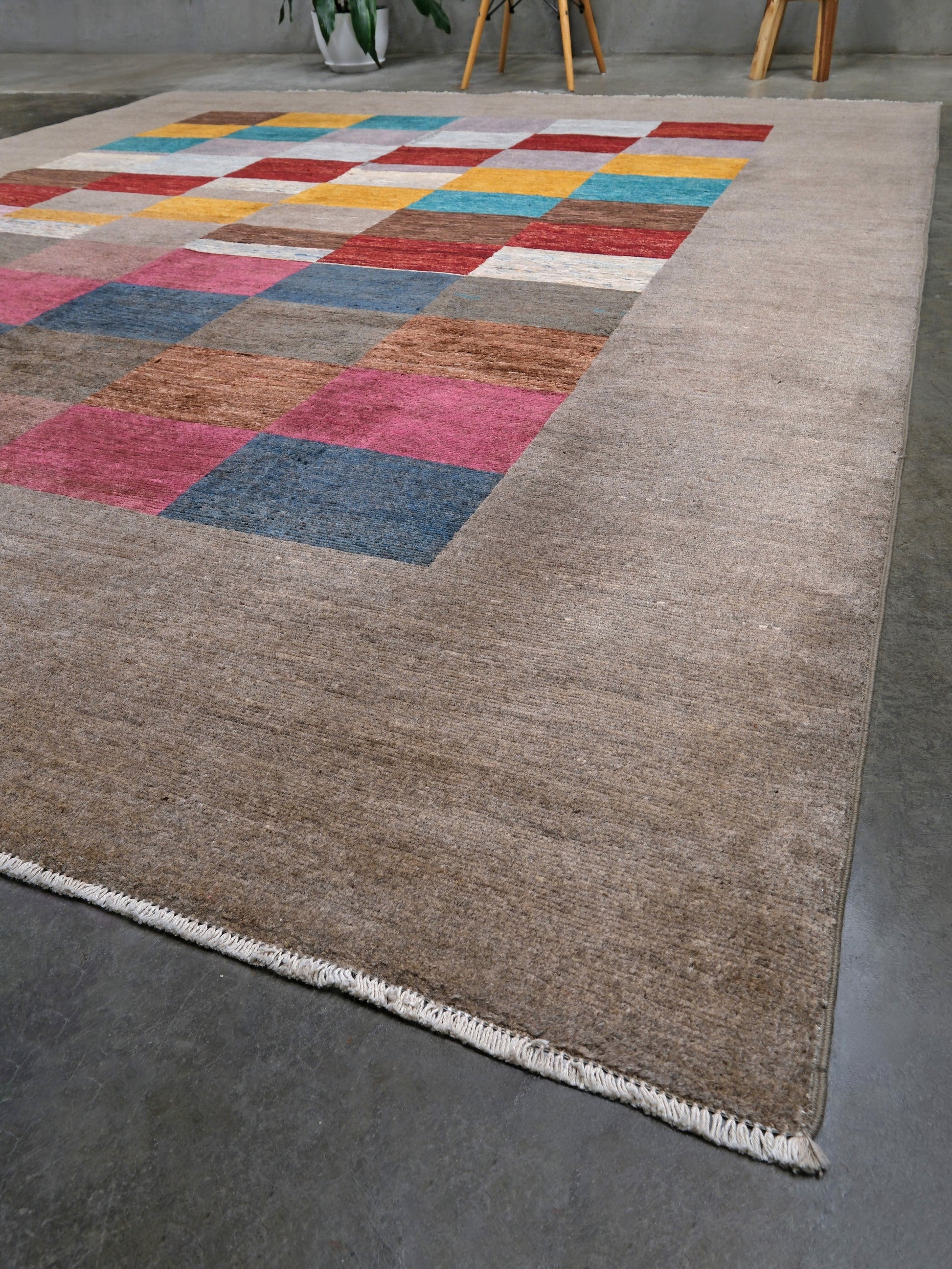 A rectangular area rug with a beige base and a colorful patchwork grid pattern in the top-left corner, shown on a gray concrete floor.