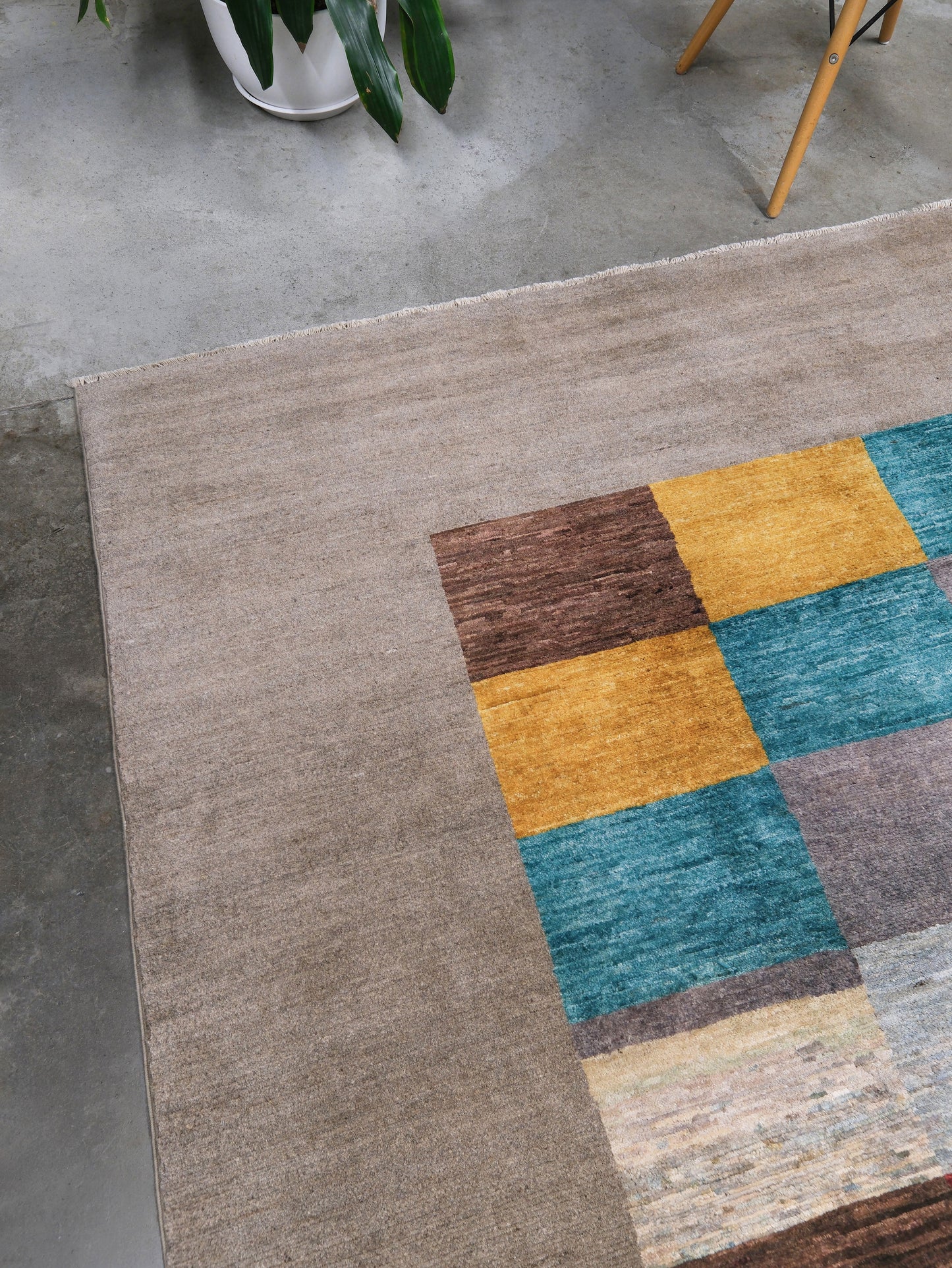 A textured area rug with a solid taupe-beige field and a geometric patchwork design in mustard yellow, teal blue, brown, and gray, shown on a concrete floor.