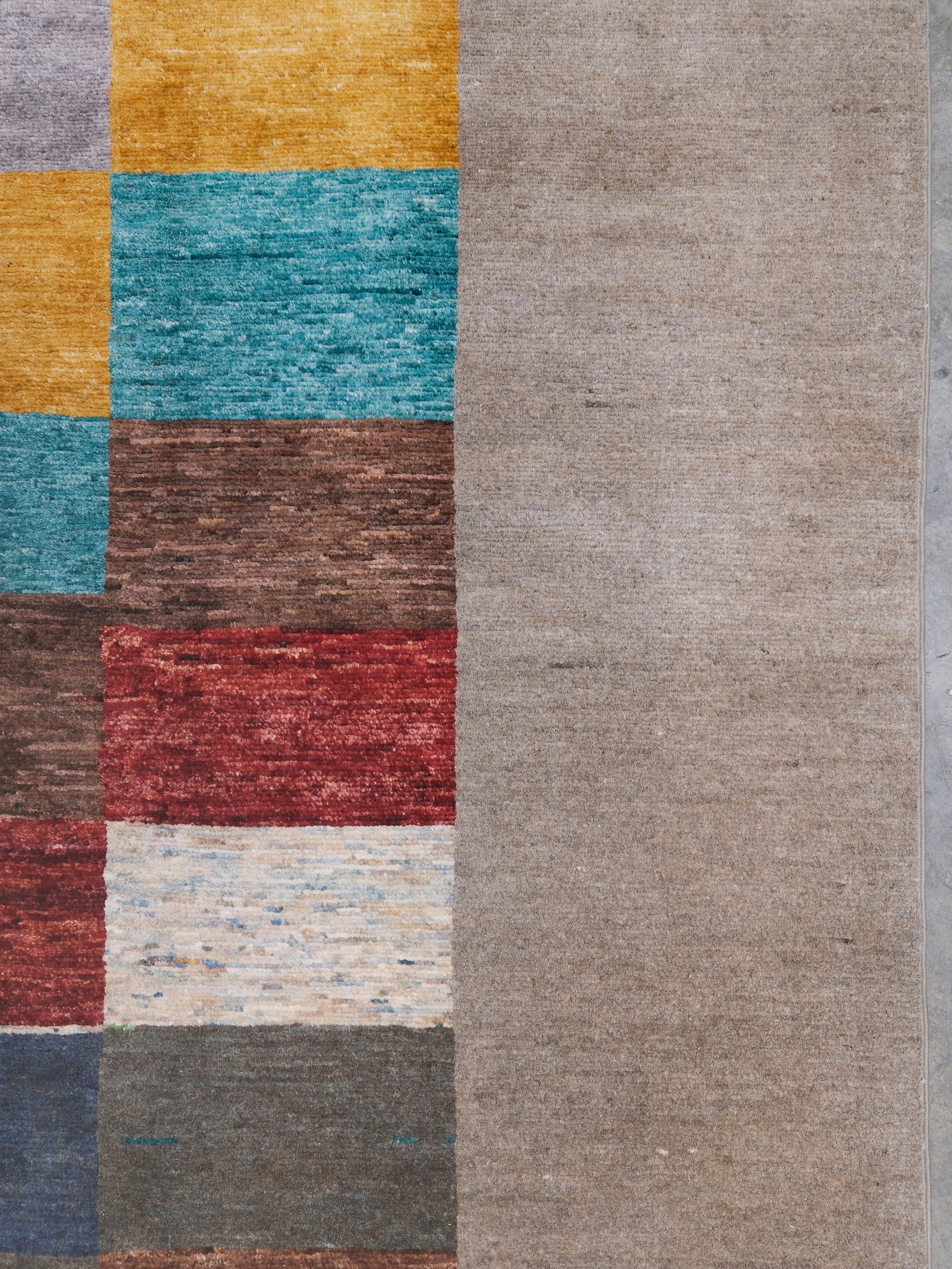 A close-up of a textured rug showing a solid taupe-beige section on the right and a multicolor block pattern on the left with yellow, teal, brown, red, and cream squares.