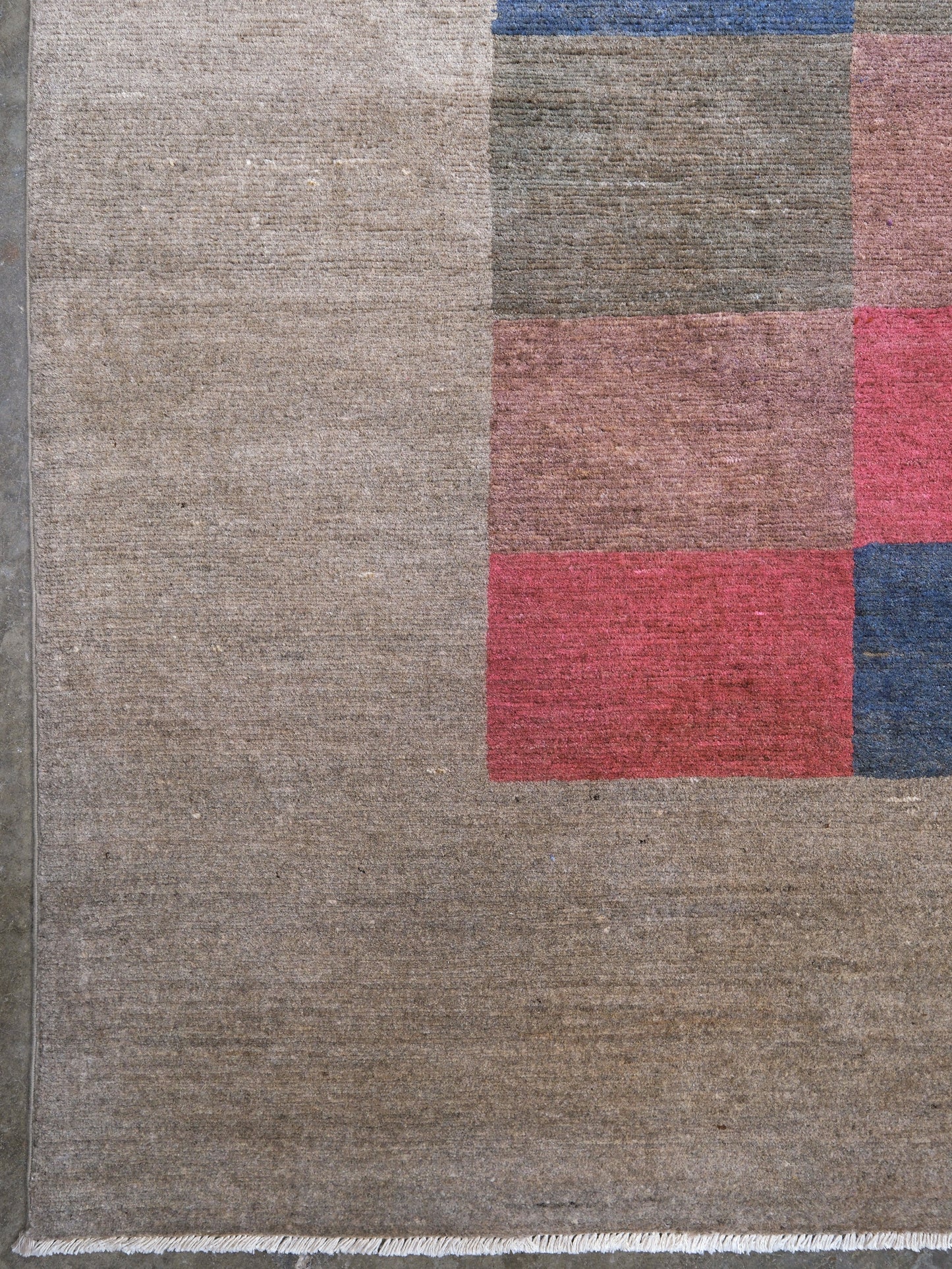 Close-up of a woven rug showing a large taupe-beige field with a partial geometric block pattern in muted red, gray-brown, and blue.