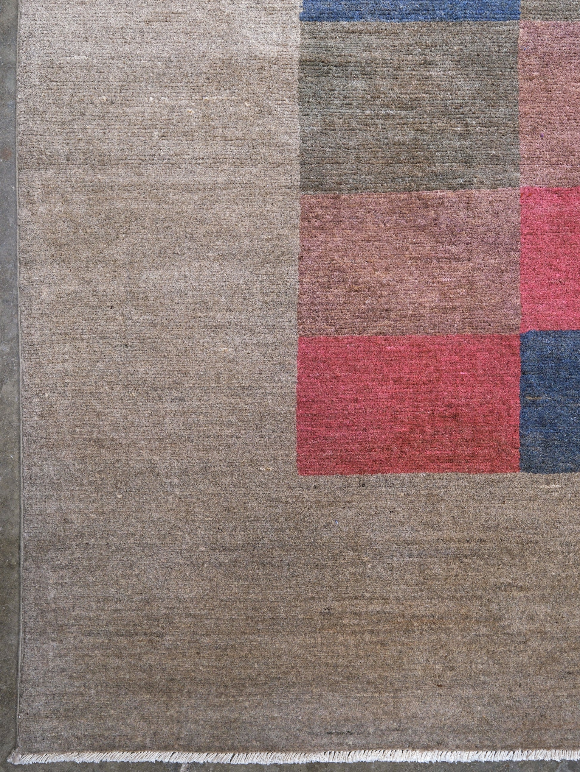 Close-up of a woven rug showing a large taupe-beige field with a partial geometric block pattern in muted red, gray-brown, and blue.