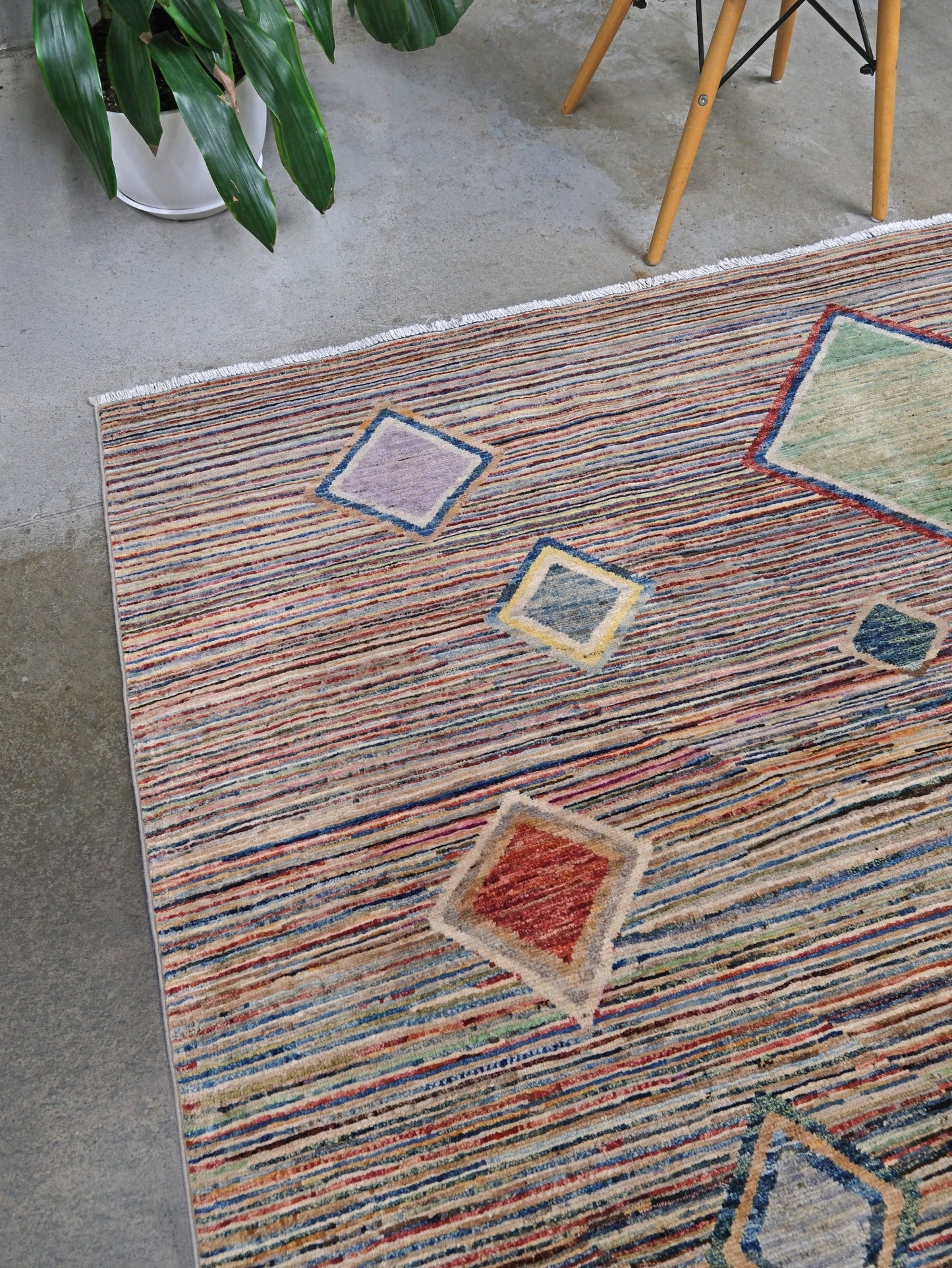 A close-up of a textured area rug with a multicolor striped pattern and geometric diamond motifs, styled on a concrete floor next to a potted plant and wooden chair legs.