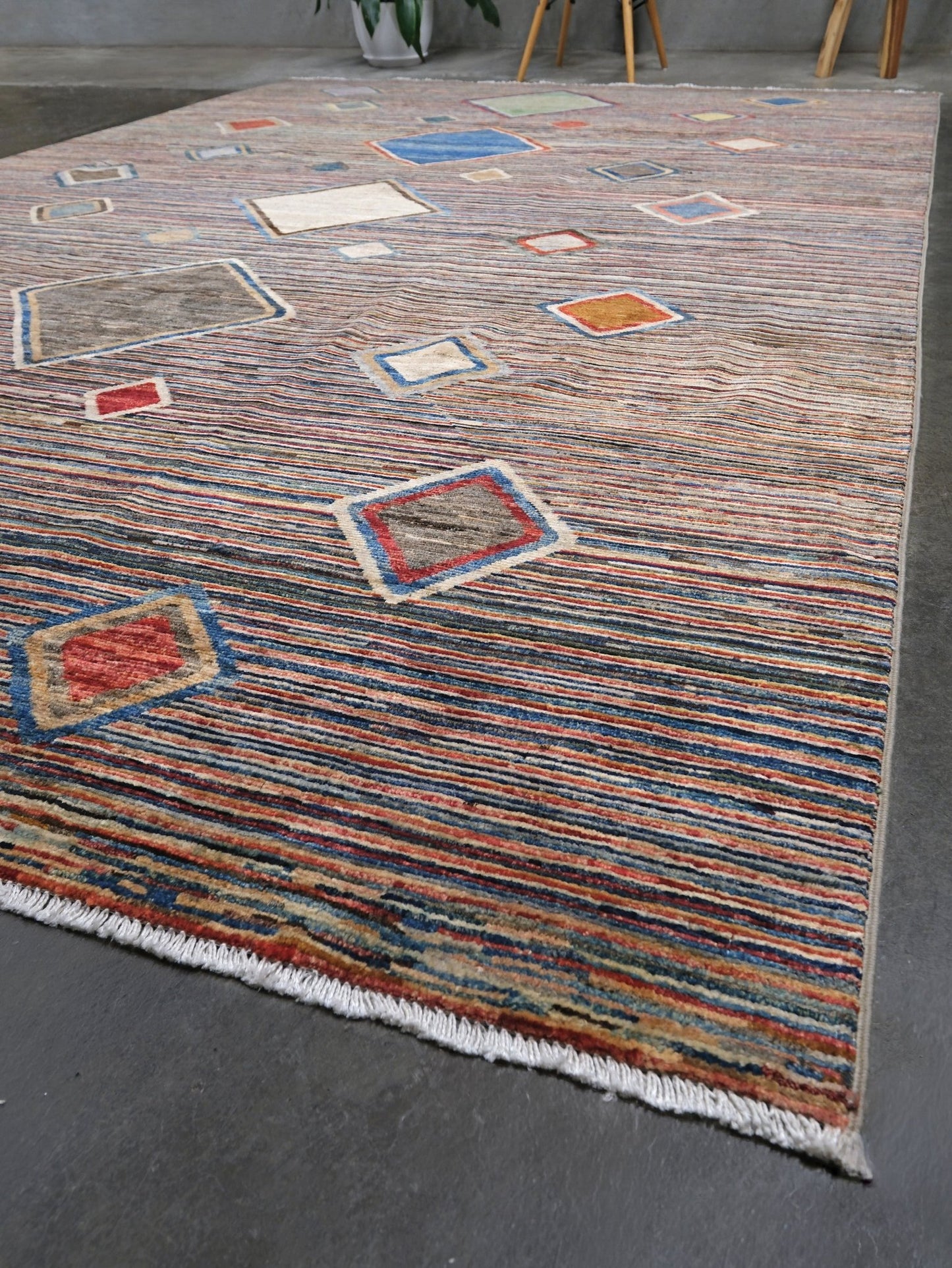 A rectangular area rug with a textured, hand-woven Moroccan style. The rug features a dominant dusty blue-gray base with numerous small, colorful geometric diamond and square motifs in red, orange, blue, beige, and green, finished with a white fringe on one end.