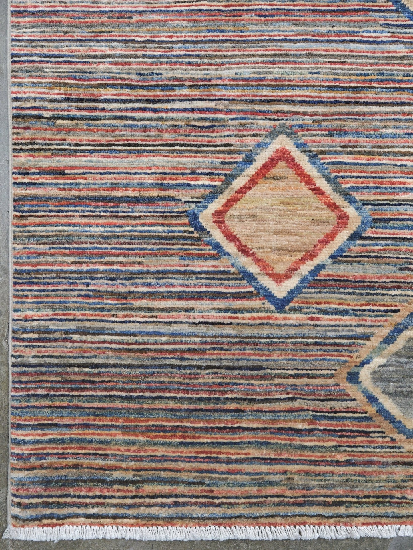A close-up of a woven rug with horizontal multicolor stripes in blue, red, beige, and gray, featuring a central diamond motif with a red border and blue outline.