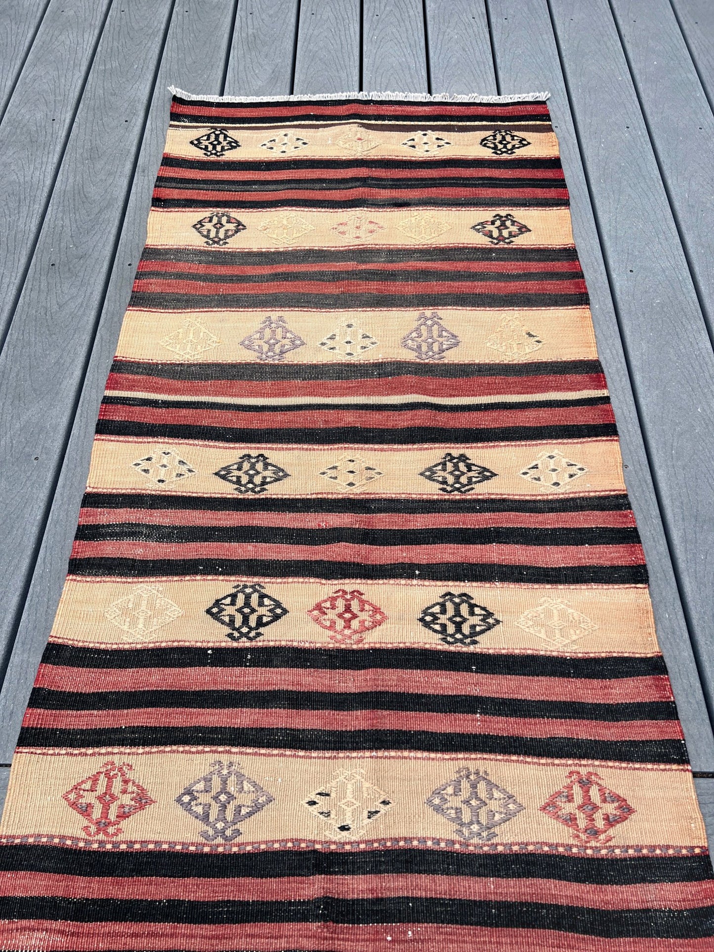 A long kilim runner rug with horizontal stripes and diamond motifs, laid out on a gray wooden deck.
