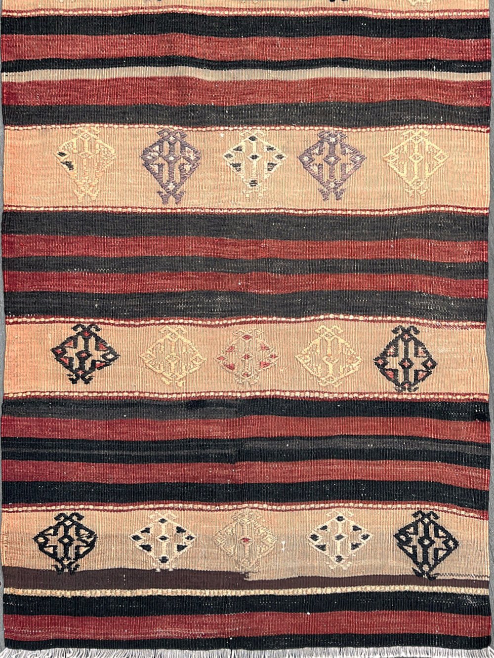 A flat-laid kilim rug with horizontal stripes in red, black, and beige, featuring diamond and geometric motifs in the center bands.