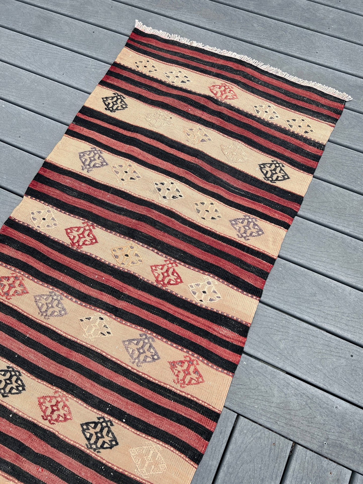 A kilim runner rug with horizontal stripes and diamond motifs, laid out diagonally on a gray wooden deck.