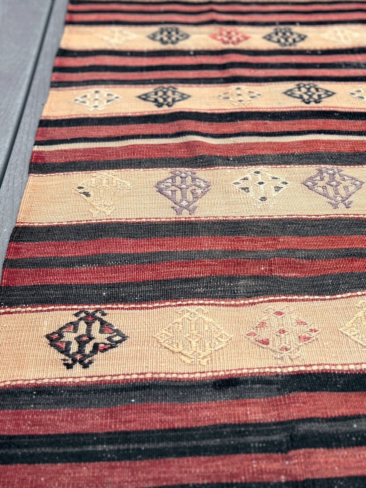 A close-up of a kilim rug featuring horizontal stripes in red, black, and beige with geometric diamond patterns.