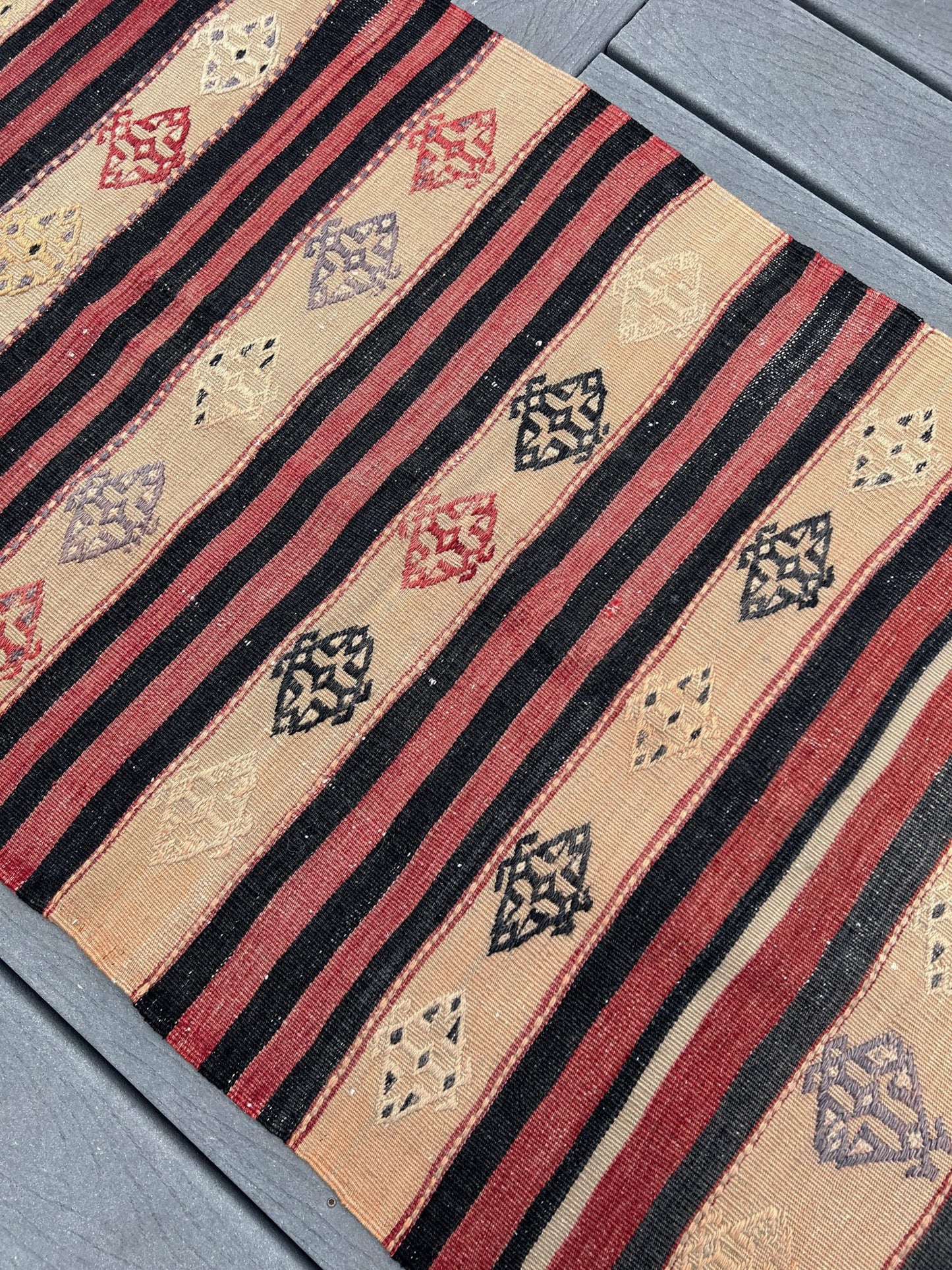 A close-up of a kilim textile runner laid diagonally on gray wooden decking. The runner features vertical stripes in black and muted red on a warm beige base, interspersed with small diamond motifs in black, red, and beige.