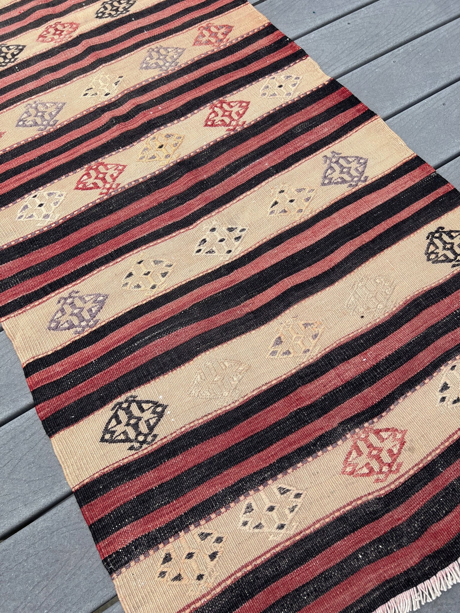 A kilim-style woven rug with horizontal stripes and diamond motifs, laid out diagonally on gray wooden deck planks.
