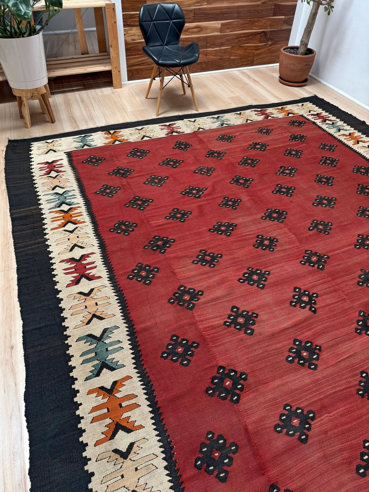 A large rectangular area rug with a red field, black geometric motifs, and a cream border with multicolor accents, shown in a modern room setting.
