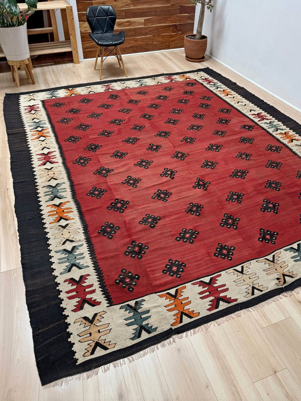 A rectangular area rug with a red field, black border, and multicolor geometric motifs, shown on a light wood floor in a modern room setting.