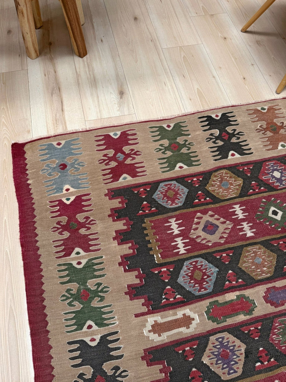 A close-up of a traditional kilim area rug with geometric patterns in red, green, black, and beige, laid out on a light wood floor next to wooden furniture legs.