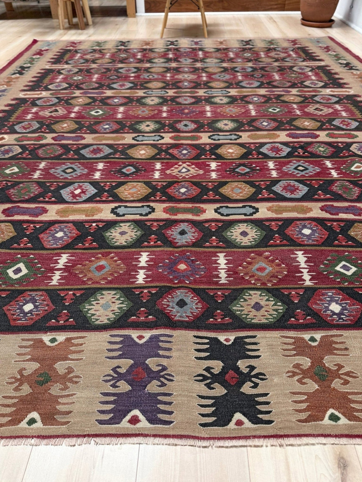 A large rectangular area rug with a complex geometric tribal pattern in red, beige, black, green, and purple, laid out on a light wood floor.