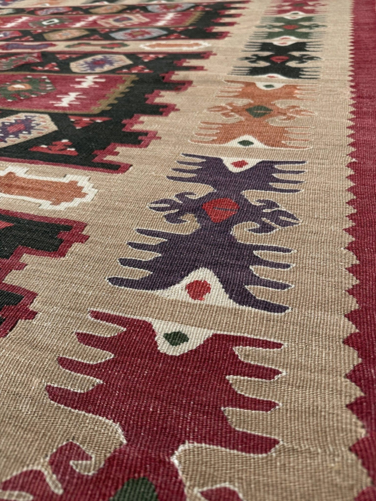Close-up of a woven kilim rug showing a beige base with bold geometric and tribal motifs in red, dark green, purple, and brown.