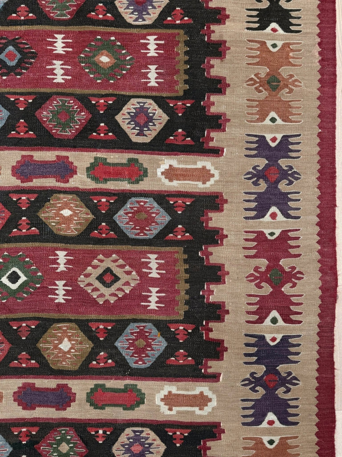 A close-up of a woven kilim rug showing a detailed geometric pattern with horizontal bands of red, black, beige, and multicolor motifs.