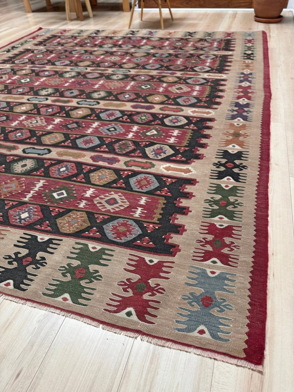 A rectangular kilim-style area rug with a complex geometric and tribal pattern, laid out on a light-colored wooden floor. The rug features a beige base with red, black, green, and blue patterns, and is shown from an elevated perspective.
