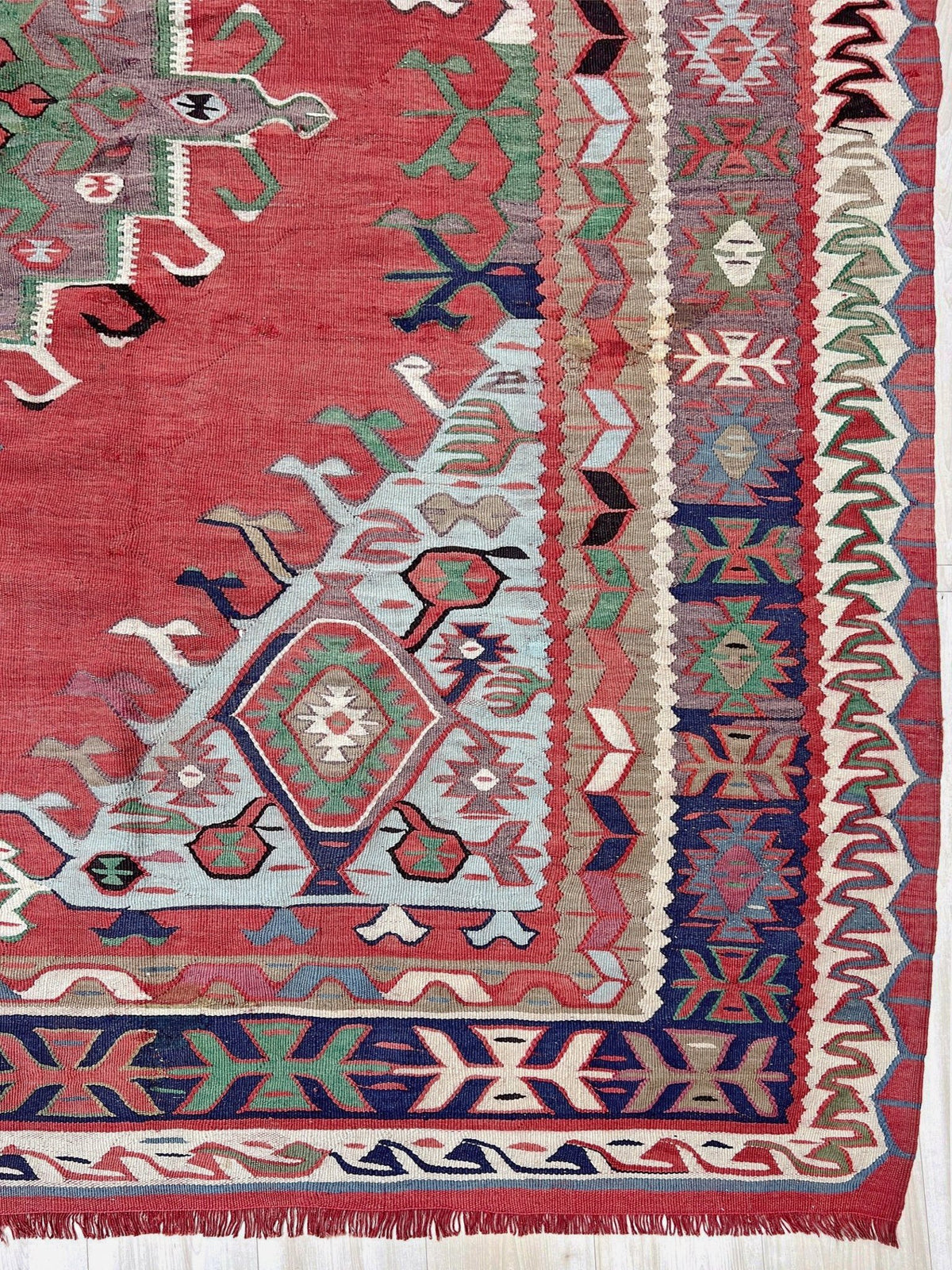 A close-up of a patterned rug with a red field, cream, blue, green, and brown geometric motifs, and fringed edges.