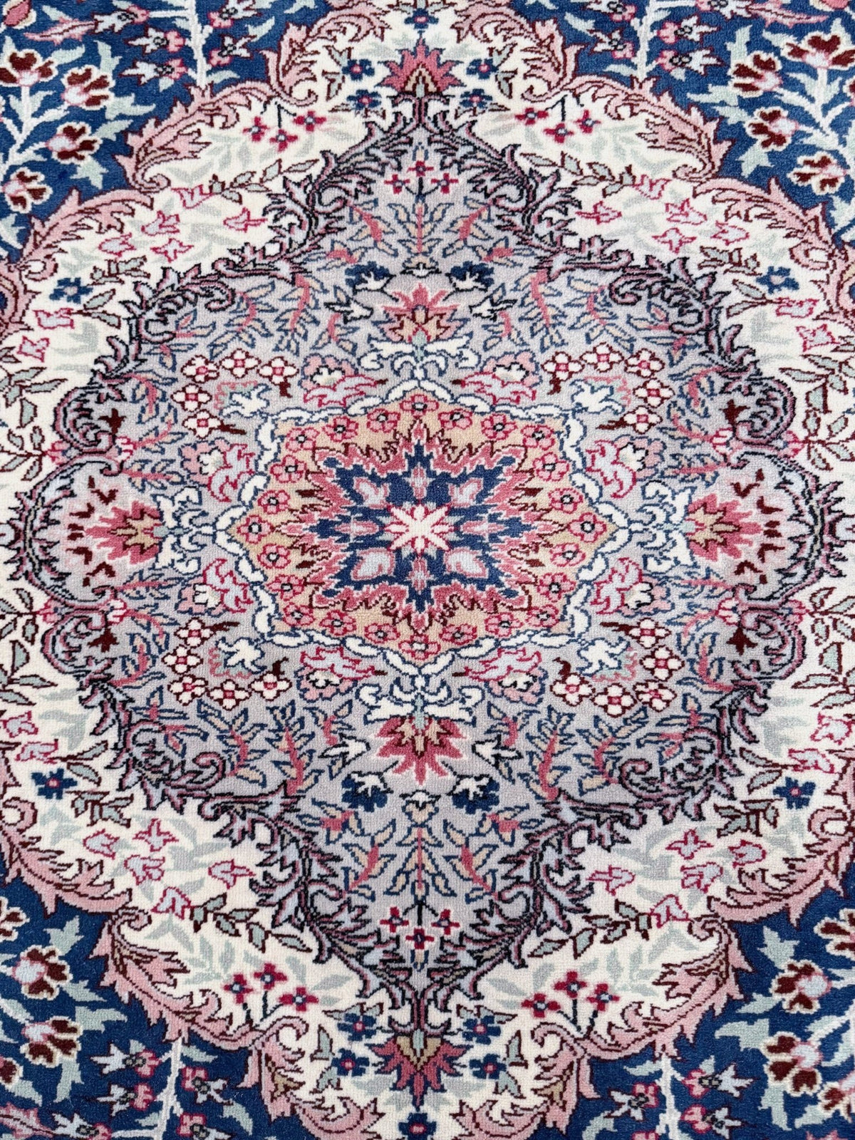 A close-up of an ornate traditional patterned rug with a central medallion, featuring cream, dusty pink, slate blue, and navy floral and vine motifs.