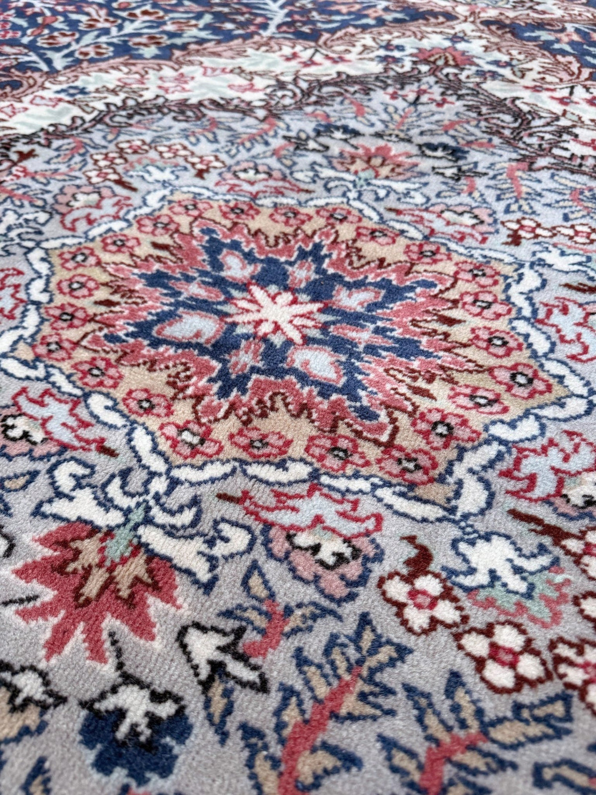 A close-up of a traditional patterned rug featuring a central medallion in shades of dusty pink, navy blue, red, and cream on a light gray base. Blue vintage Turkish rug with deep blue field,  floral medallion, handmade wool 7x9 from Indigo Rugs Burlingame Bay Area.