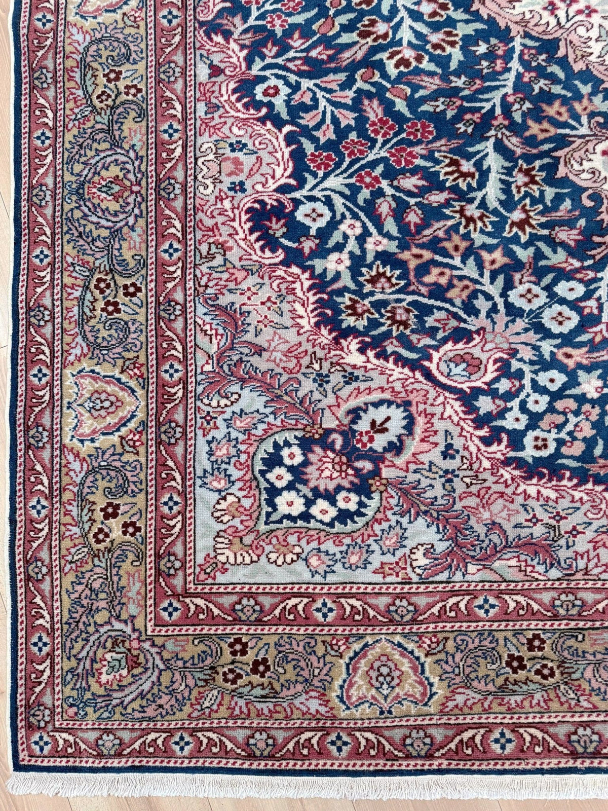 A close-up of an ornate Persian-style area rug showing the corner and border details. The design features a deep navy blue field with intricate floral motifs in red, pink, beige, and white, surrounded by a detailed multi-colored border.