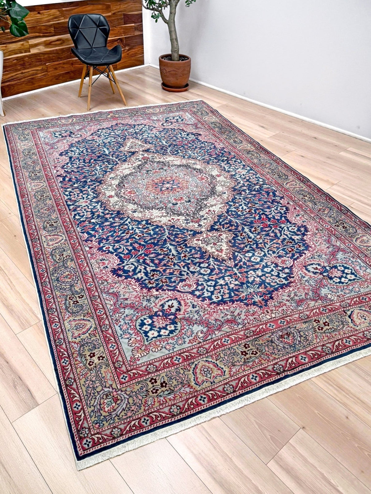 A traditional oriental area rug with an intricate floral pattern in navy blue, red, beige, and gray, displayed on a light wood floor in a modern room setting. Blue vintage Turkish rug with deep blue field,  floral medallion, handmade wool 7x9 from Indigo Rugs Burlingame Bay Area.
