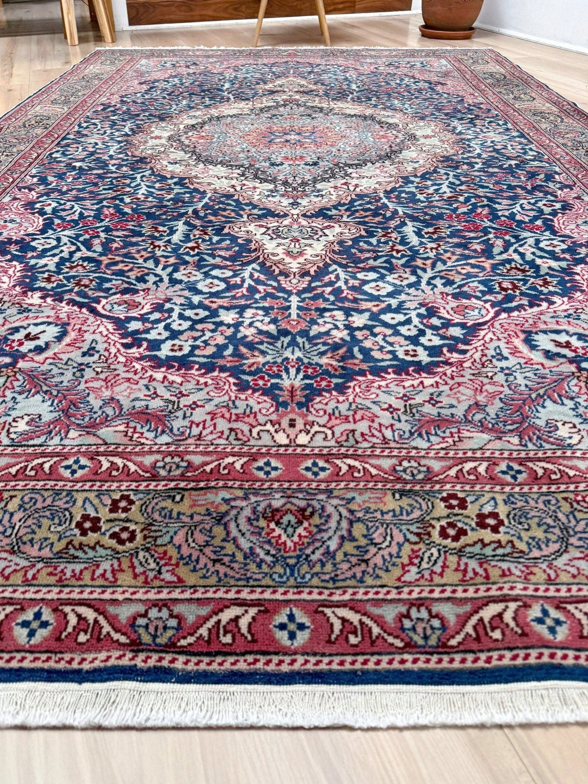 A traditional Persian-style area rug with a complex floral and medallion pattern, featuring a deep blue field, red and pink borders, and cream accents, shown on a light wood floor. Blue vintage Turkish rug with deep blue field,  floral medallion, handmade wool 7x9 from Indigo Rugs Burlingame Bay Area.