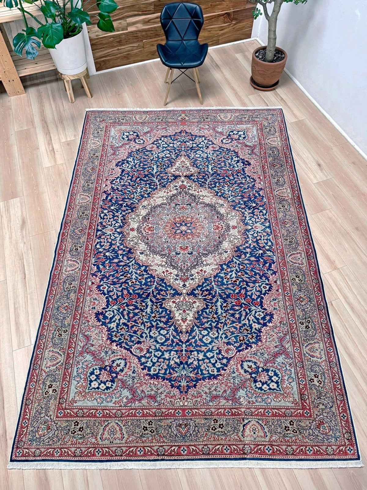 A traditional Persian-style area rug with a complex floral and medallion pattern in navy blue, red, beige, and cream, shown laid out on a light wood floor in a room setting with a chair and plants. Blue vintage Turkish rug with deep blue field,  floral medallion, handmade wool 7x9 from Indigo Rugs Burlingame Bay Area.