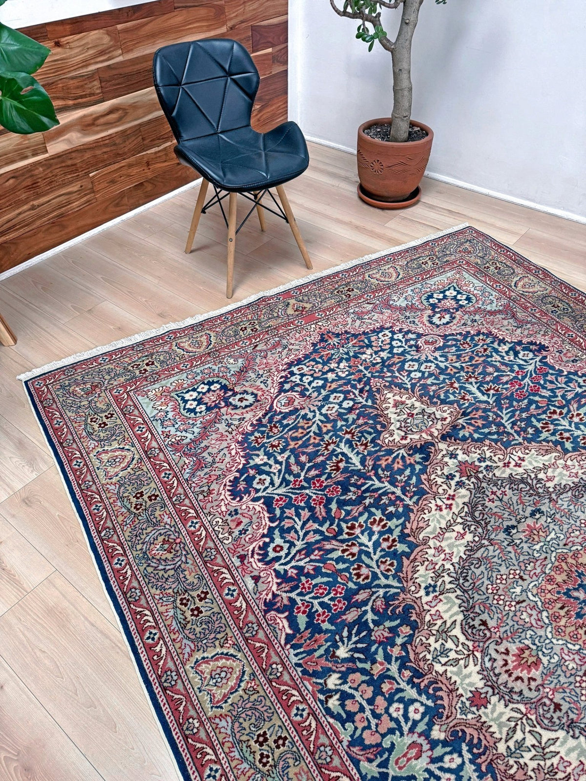 A traditional Persian-style area rug with an intricate floral pattern in navy blue, red, beige, and green, shown on a light wood floor in a room setting next to a black chair and a potted plant. Blue vintage Turkish rug with deep blue field,  floral medallion, handmade wool 7x9 from Indigo Rugs Burlingame Bay Area.