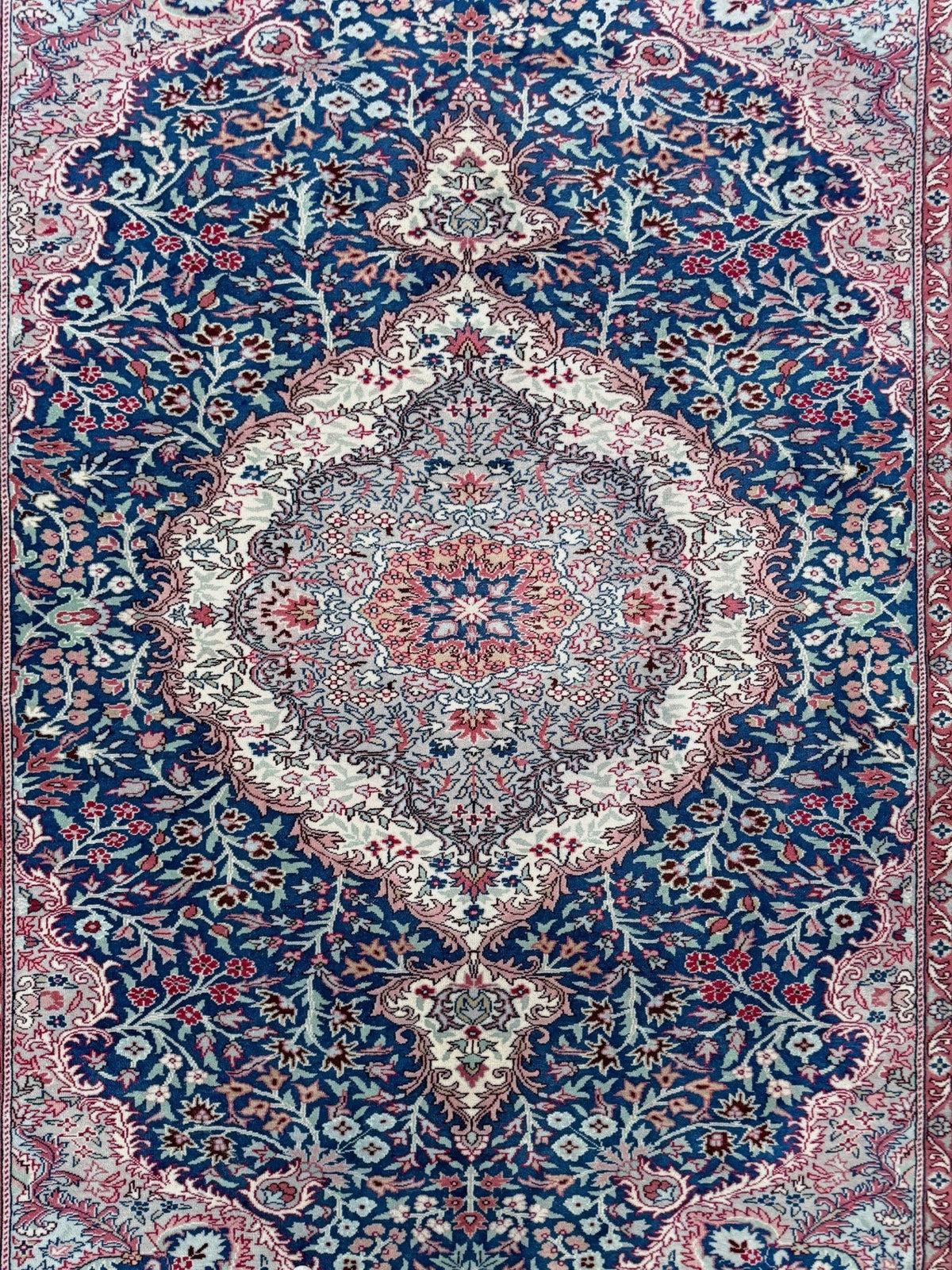 A traditional Persian-style area rug with an intricate floral pattern, featuring a large central medallion and a deep blue field with red, beige, and cream details.