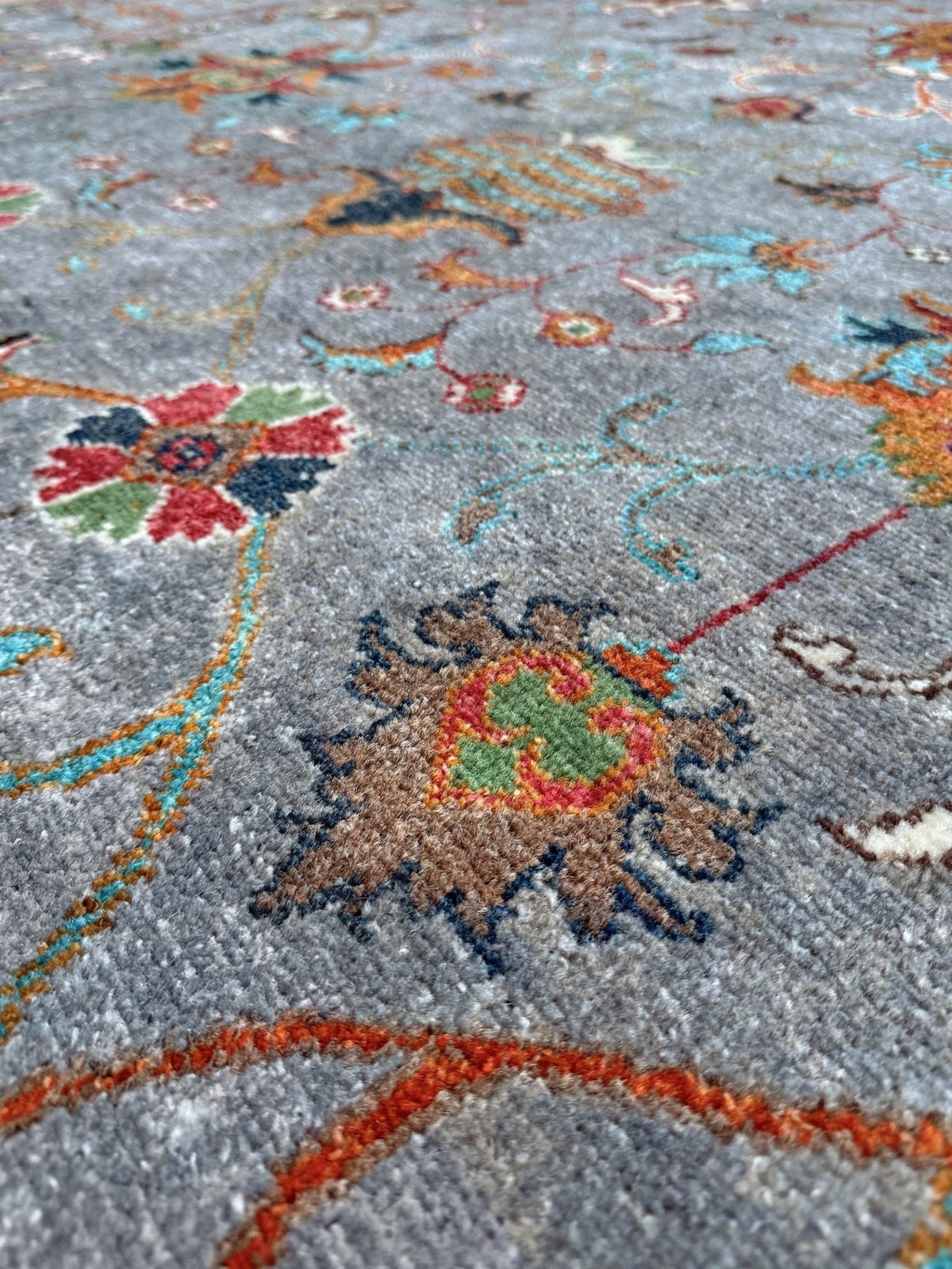 Close-up of a textured area rug with a medium gray base and a traditional floral pattern in red, green, orange, and blue. Blue gray Ziegler Turkish rug with floral accents, handmade wool 7x9 area rug from Indigo Rugs Burlingame Bay Area.