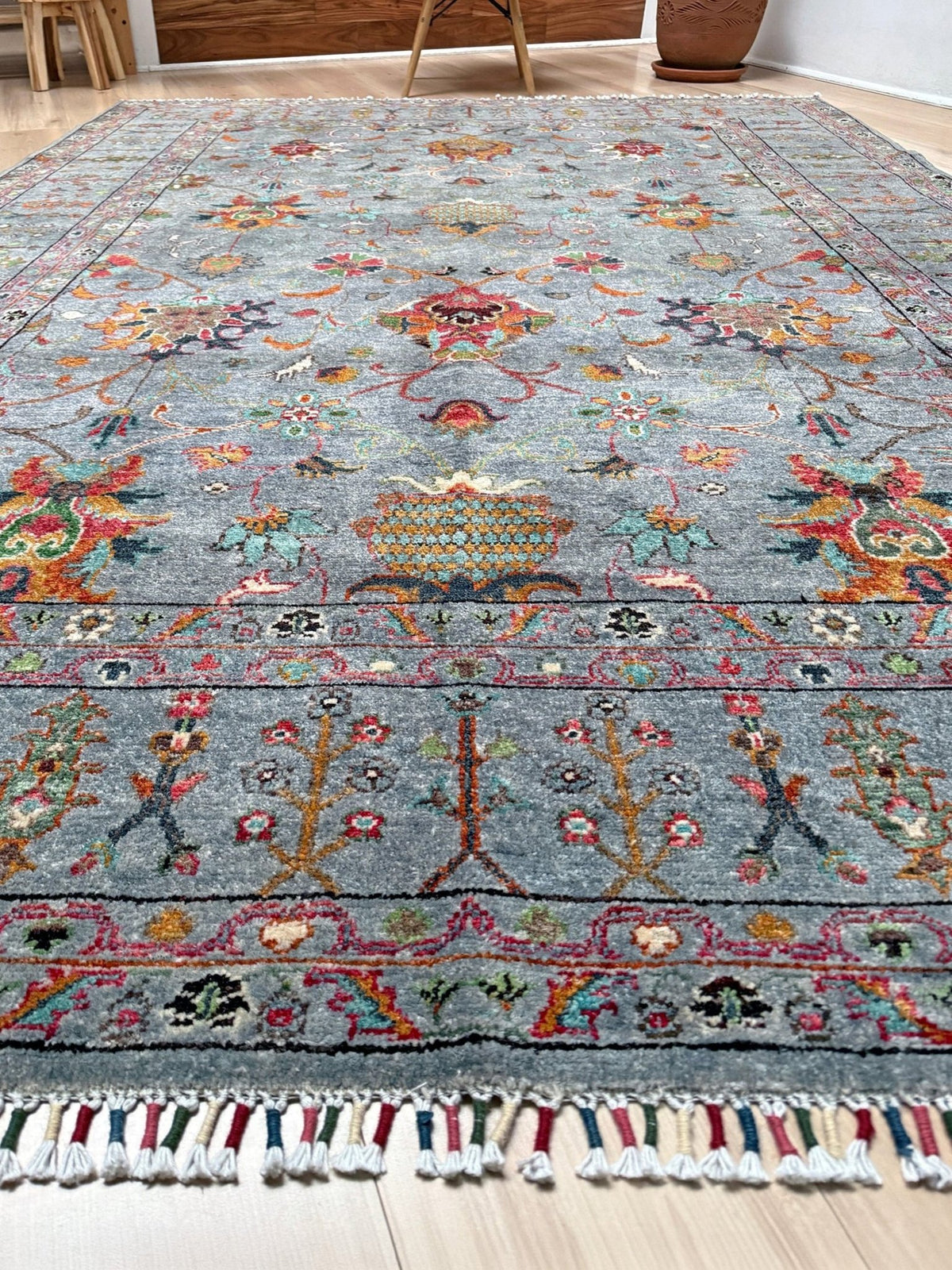 A rectangular area rug with a distressed blue-gray background and intricate multicolor floral and geometric patterns, featuring a decorative border and white tassel fringe, shown on a light wood floor. Blue gray Ziegler Turkish rug with floral accents, handmade wool 7x9 area rug from Indigo Rugs Burlingame Bay Area.