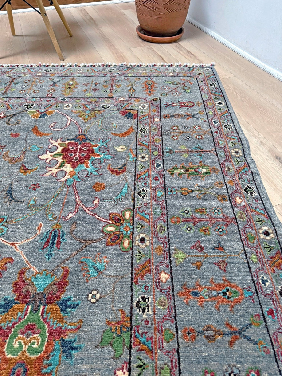 A rectangular area rug with a distressed gray base and intricate multicolor traditional patterns, shown laid out on a light wood floor in a room setting. Blue gray Ziegler Turkish rug with floral accents, handmade wool 7x9 area rug from Indigo Rugs Burlingame Bay Area.