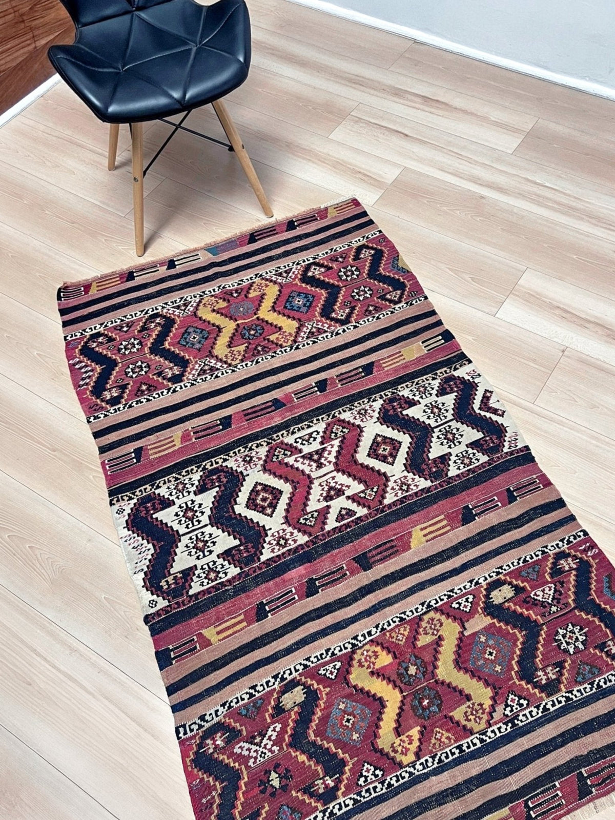 A long rectangular area rug with a traditional geometric tribal pattern in red, navy, beige, and yellow, laid out on a light wood floor next to a black leather chair.