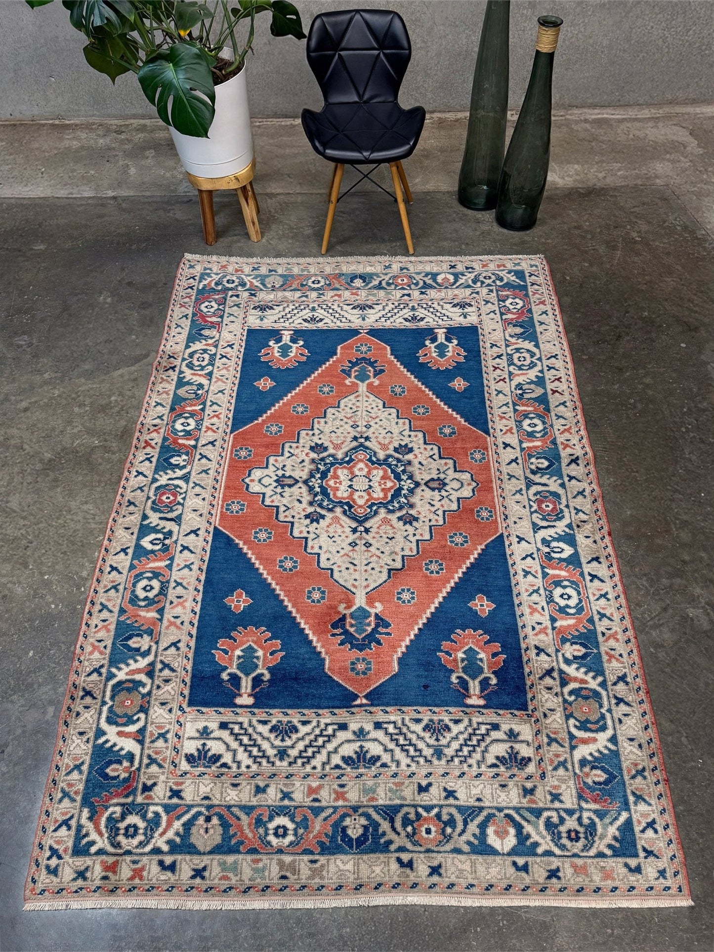 Decorative rug with geometric pattern on a concrete floor, with a chair and plants in the background. Vintage turkish taspinar rug 6x8 indigo blue, salmon red  hand knotted anatolian wool rug pastel san francisco bay area rug shop indigo rugs