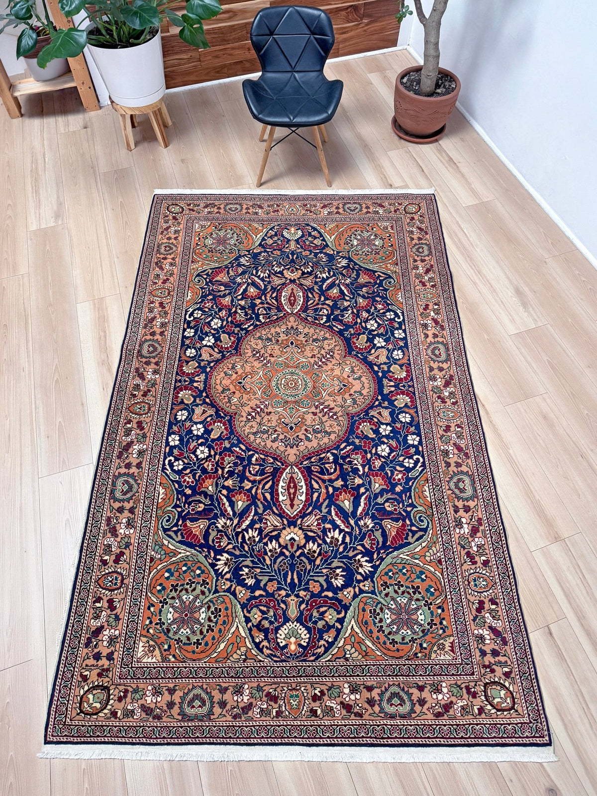 A long rectangular area rug with an ornate traditional Persian-style pattern in deep blue, rust red, beige, and green, shown on a light wood floor in a modern room setting. 5x7 Navy Kayseri Turkish rug with peach medallion, floral vines, handmade wool from Indigo Rugs Burlingame Bay Area.