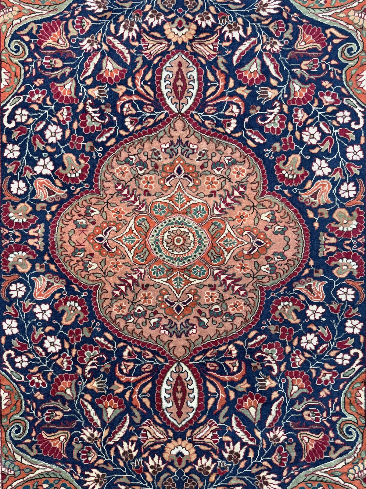 A traditional Persian-style area rug with an intricate floral and medallion pattern, featuring a dominant navy blue background with a large central peach and beige medallion surrounded by red, green, and cream details. 5x7 Navy Kayseri Turkish rug with peach medallion, floral vines, handmade wool from Indigo Rugs Burlingame Bay Area.