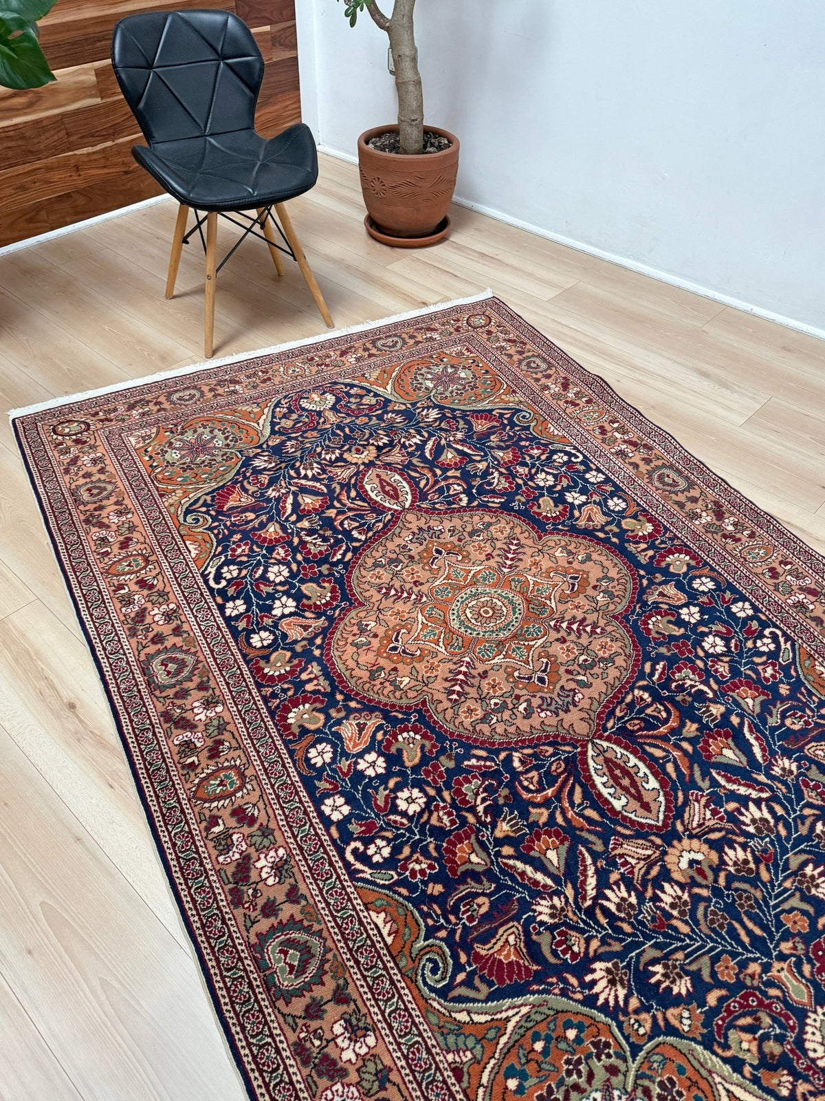 A traditional oriental-style area rug with a dark navy blue field, intricate floral motifs, and a warm rust-orange central medallion, shown on a light wood floor in a room setting with a black chair and potted plant. 5x7 Navy Kayseri Turkish rug with peach medallion, floral vines, handmade wool from Indigo Rugs Burlingame Bay Area.
