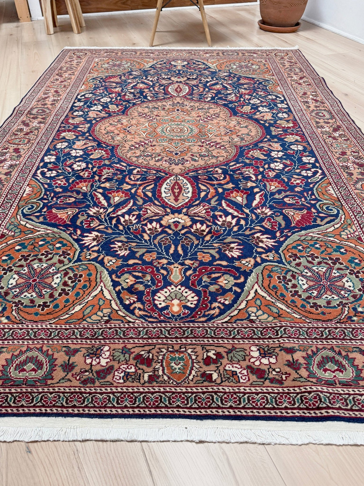 A traditional Persian-style area rug with an intricate floral and medallion pattern, displayed on a light wood floor in a room setting. 5x7 Navy Kayseri Turkish rug with peach medallion, floral vines, handmade wool from Indigo Rugs Burlingame Bay Area.