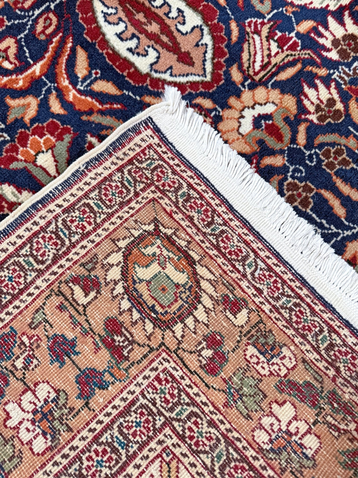 Close-up of a patterned area rug corner with white fringe, showing a beige field with red, blue, and green floral motifs, layered over another dark blue patterned rug. 5x7 Navy Kayseri Turkish rug with peach medallion, floral vines, handmade wool from Indigo Rugs Burlingame Bay Area.